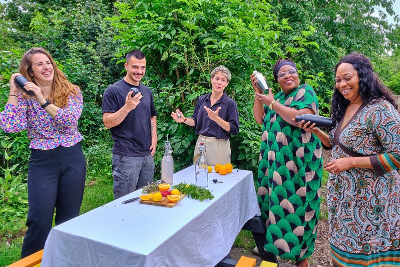 Five people standing outdoors around a table with plants, fruits, and bottles, smiling and talking. The scene appears to be a social gathering or workshop in a lush green garden.