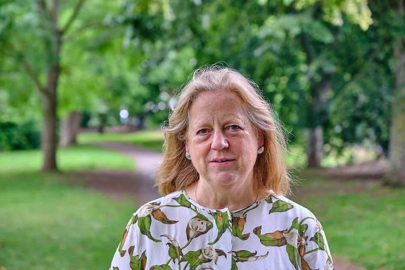 Portrait of a middle-aged woman outdoors in a park with trees and a walking path in the background.