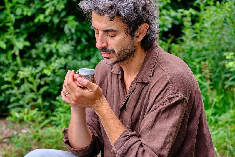 A man with gray, curly hair and a beard, wearing a brown shirt, is holding and smelling a small cup outdoors surrounded by green foliage.