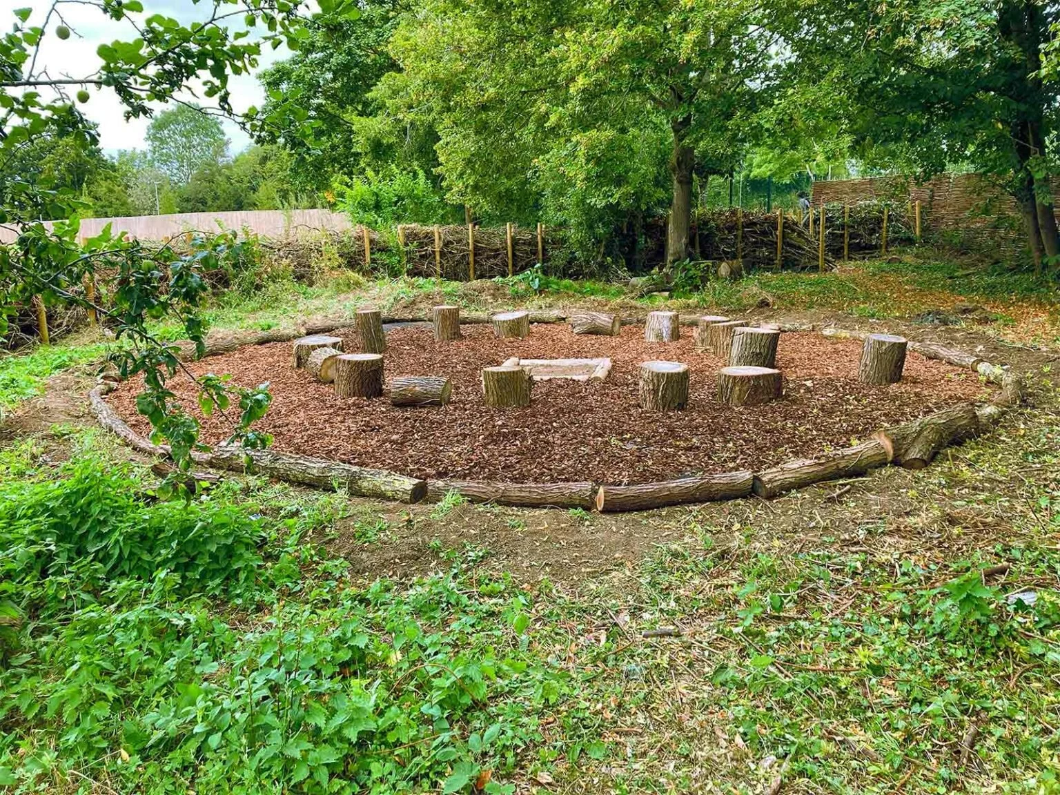 A circle of wooden logs with a ground cover of wood mulch, surrounded by green trees and bushes, with a chain-link fence in the background.