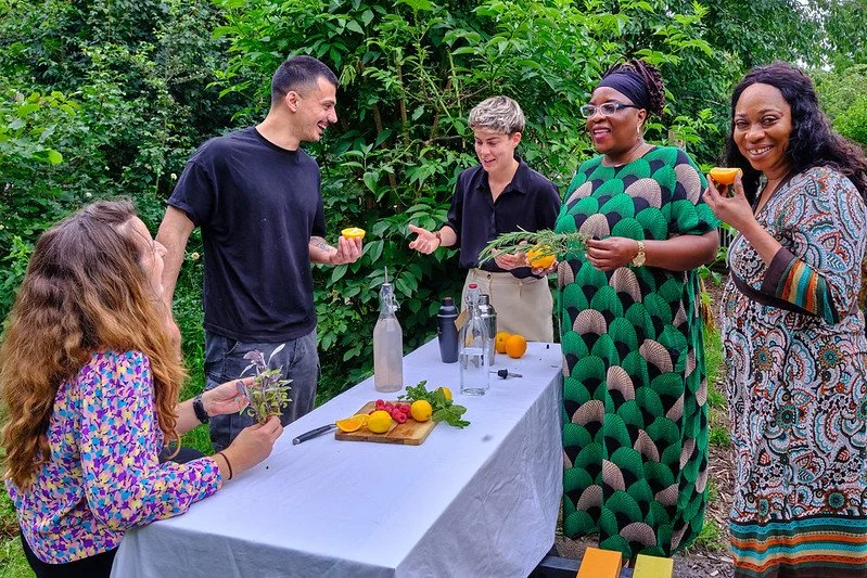 Group of five diverse women and men outdoors around a table, preparing drinks with fresh herbs and citrus, smiling and chatting.