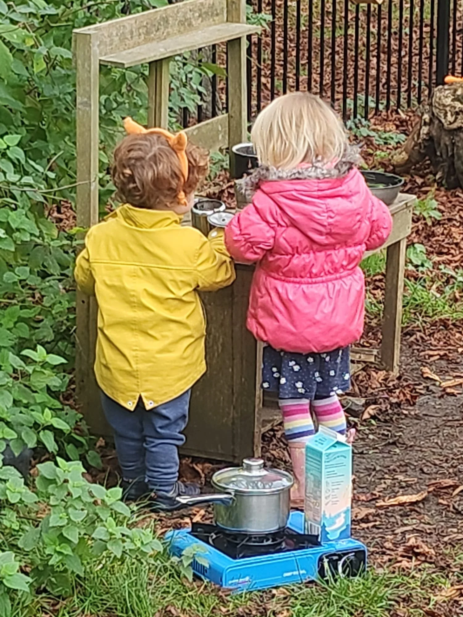 Two children, one with blonde hair wearing a pink coat and colorful rain boots and the other with brown hair wearing a yellow jacket, are standing outdoors by a wooden table with pots and utensils, surrounded by green foliage and fallen leaves.