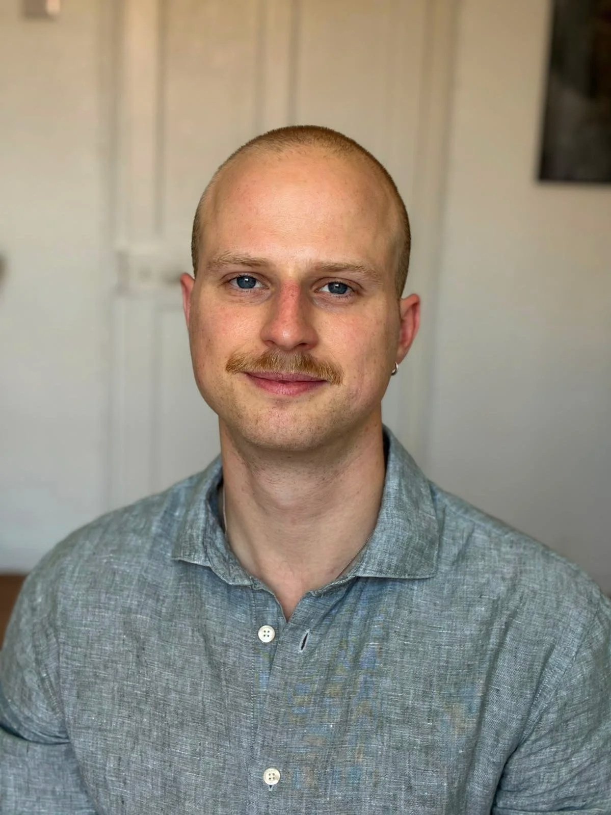 A young man with a bald head, blue eyes, and a light mustache, smiling softly, wearing a gray button-up shirt, indoors with a neutral background.