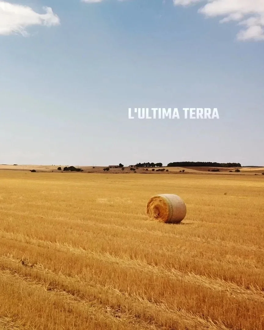 Campo con un panal di paglia rotolato, sotto un cielo azzurro con alcune nuvole, con la scritta "L'ULTIMA TERRA" in primo piano.