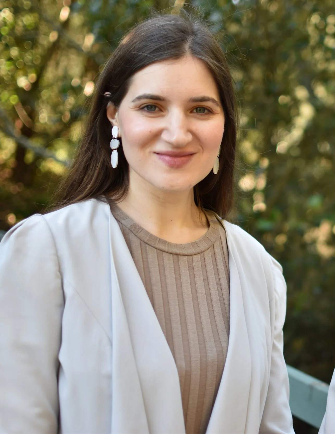 A young woman with long brown hair, wearing white earrings, a beige ribbed top, and a white blazer, stands outdoors with blurred trees in the background.