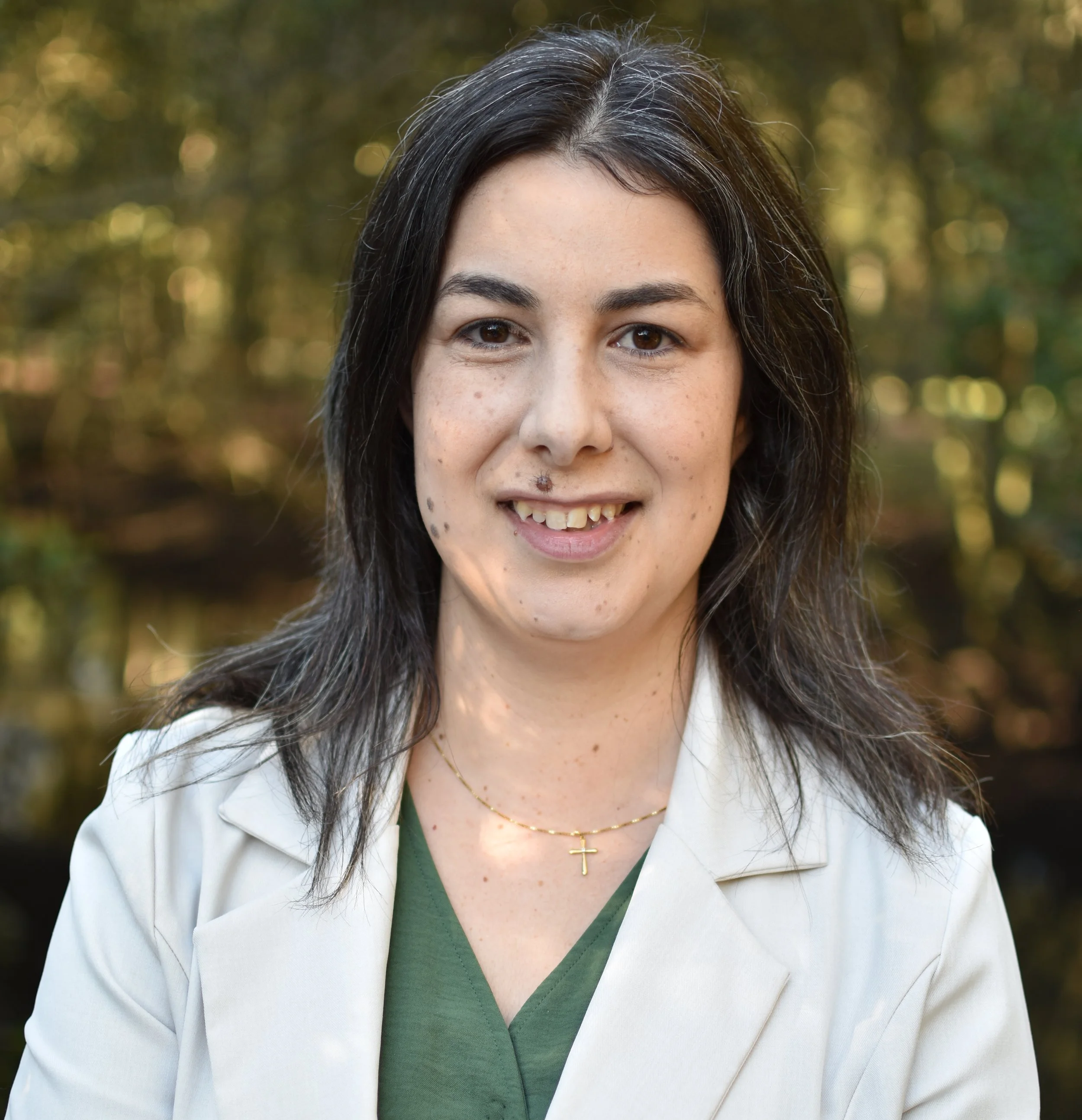 A person with dark hair, smiling, wearing a white blazer over a green shirt, in an outdoor setting with blurred greenery in the background.