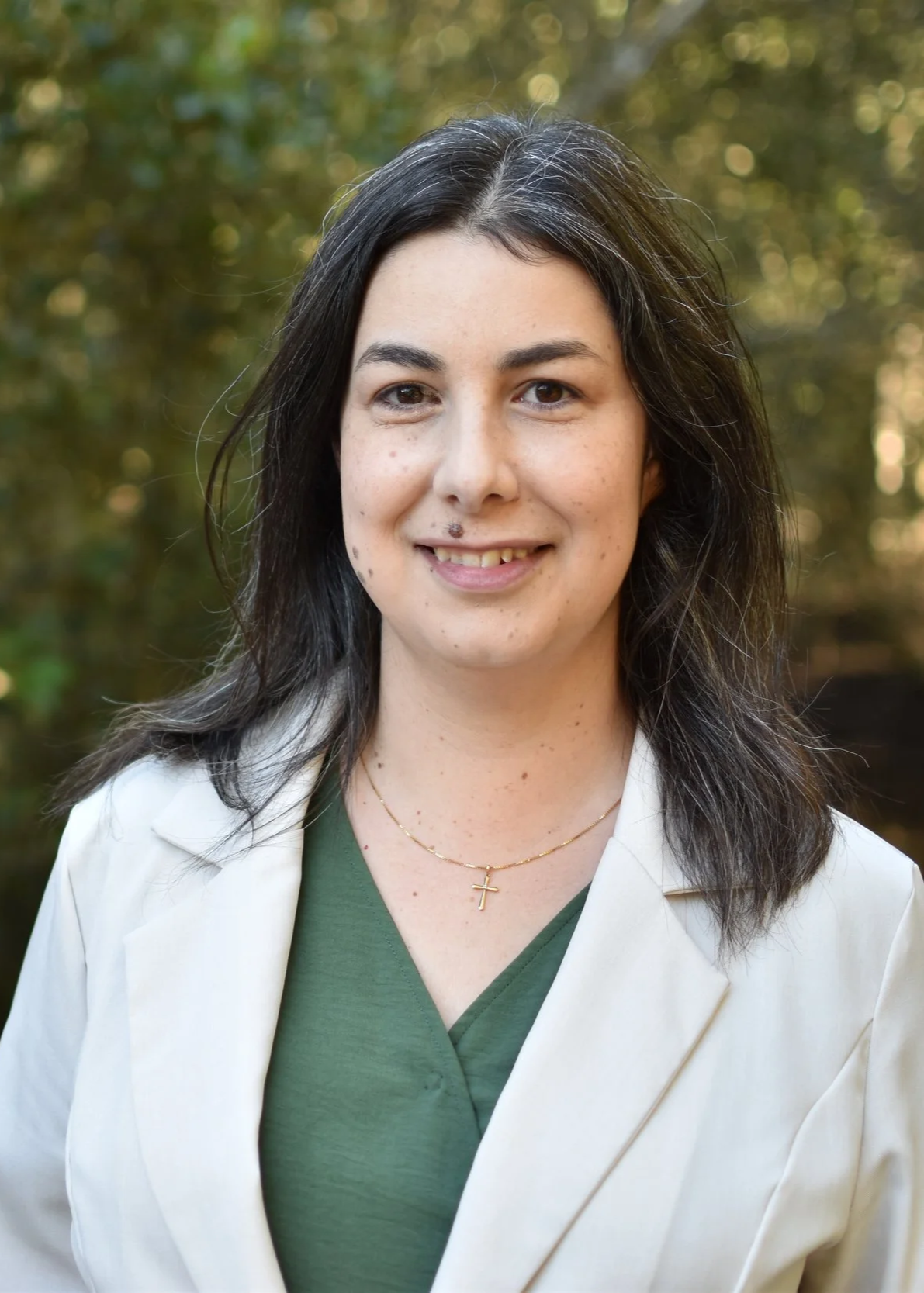 A woman with dark brown hair, wearing a white blazer, green top, and a gold cross necklace, standing outdoors with a blurred green background, smiling slightly.