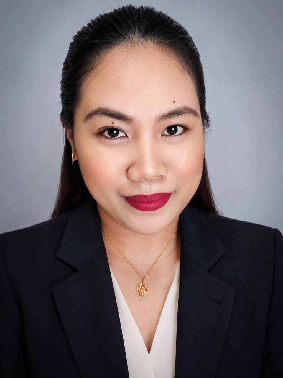 A woman with long dark hair, wearing a black blazer over a white top, gold necklace, and red lipstick, poses for a professional headshot against a gray background.