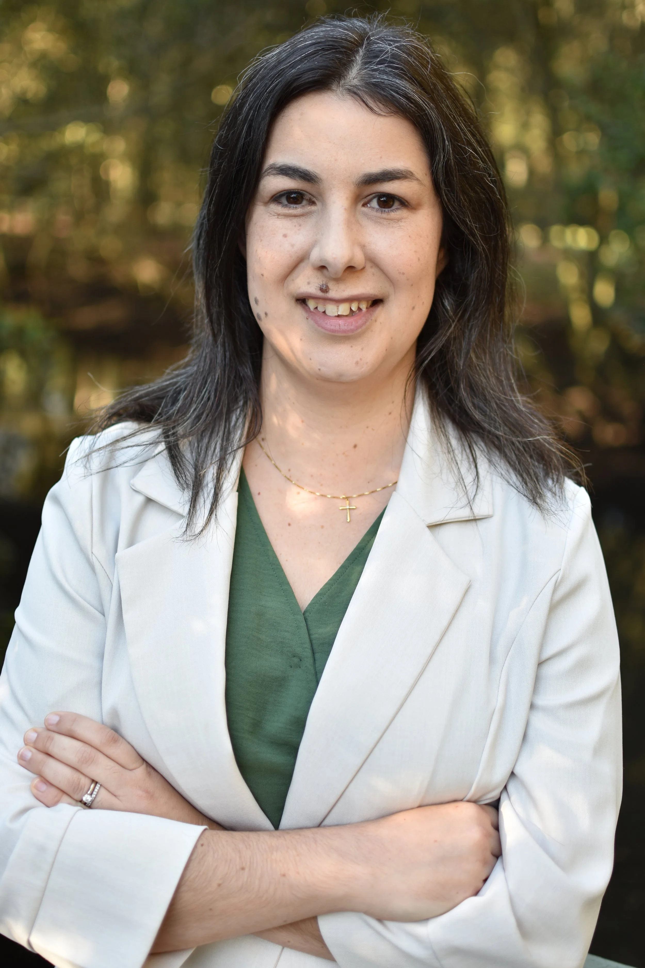Portrait of a woman with dark hair, wearing a white coat and a green top, standing outdoors with a blurred forest background.