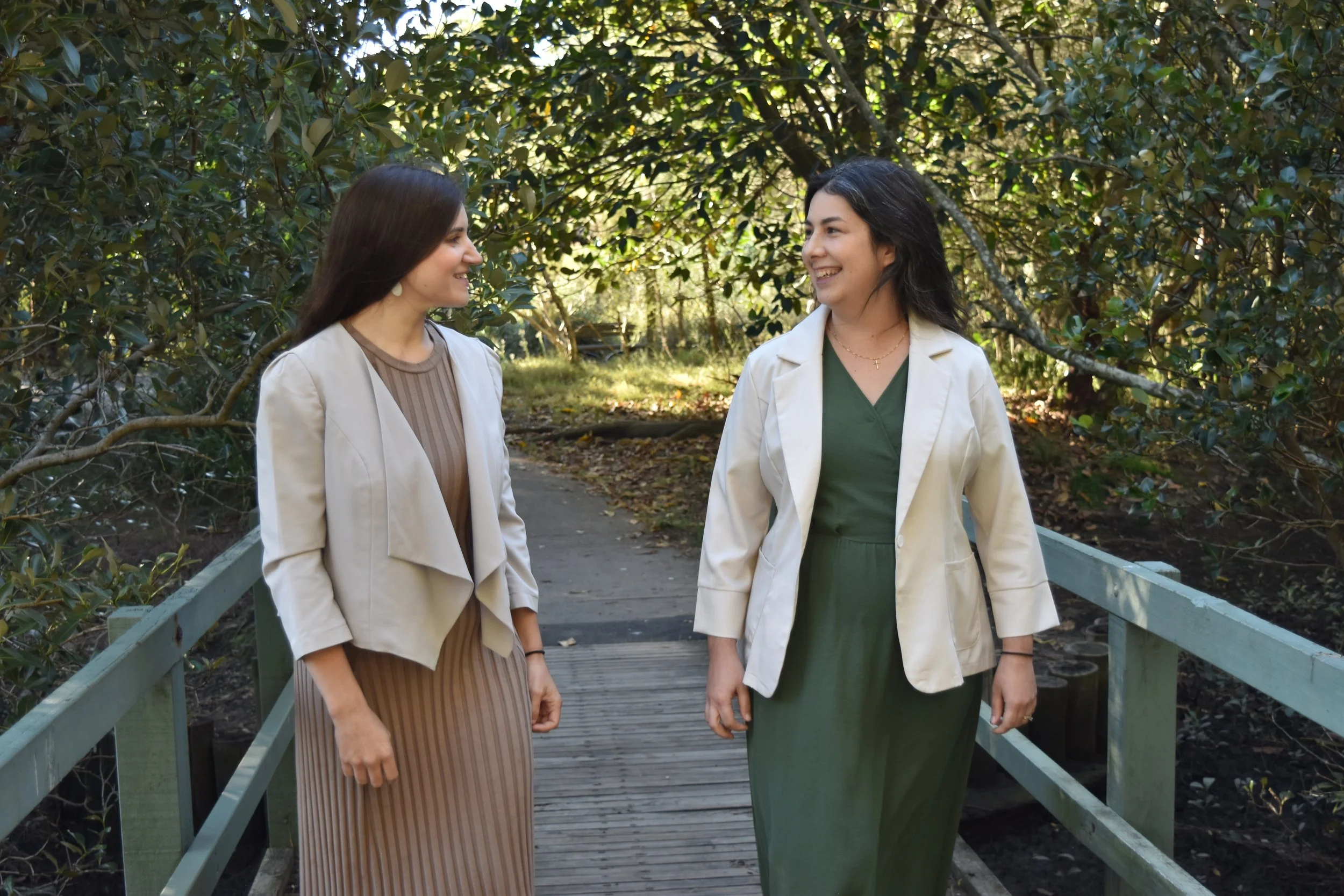 Two women dressed in beige and green walking outdoors on a wooden bridge surrounded by trees and greenery, smiling and looking at each other.