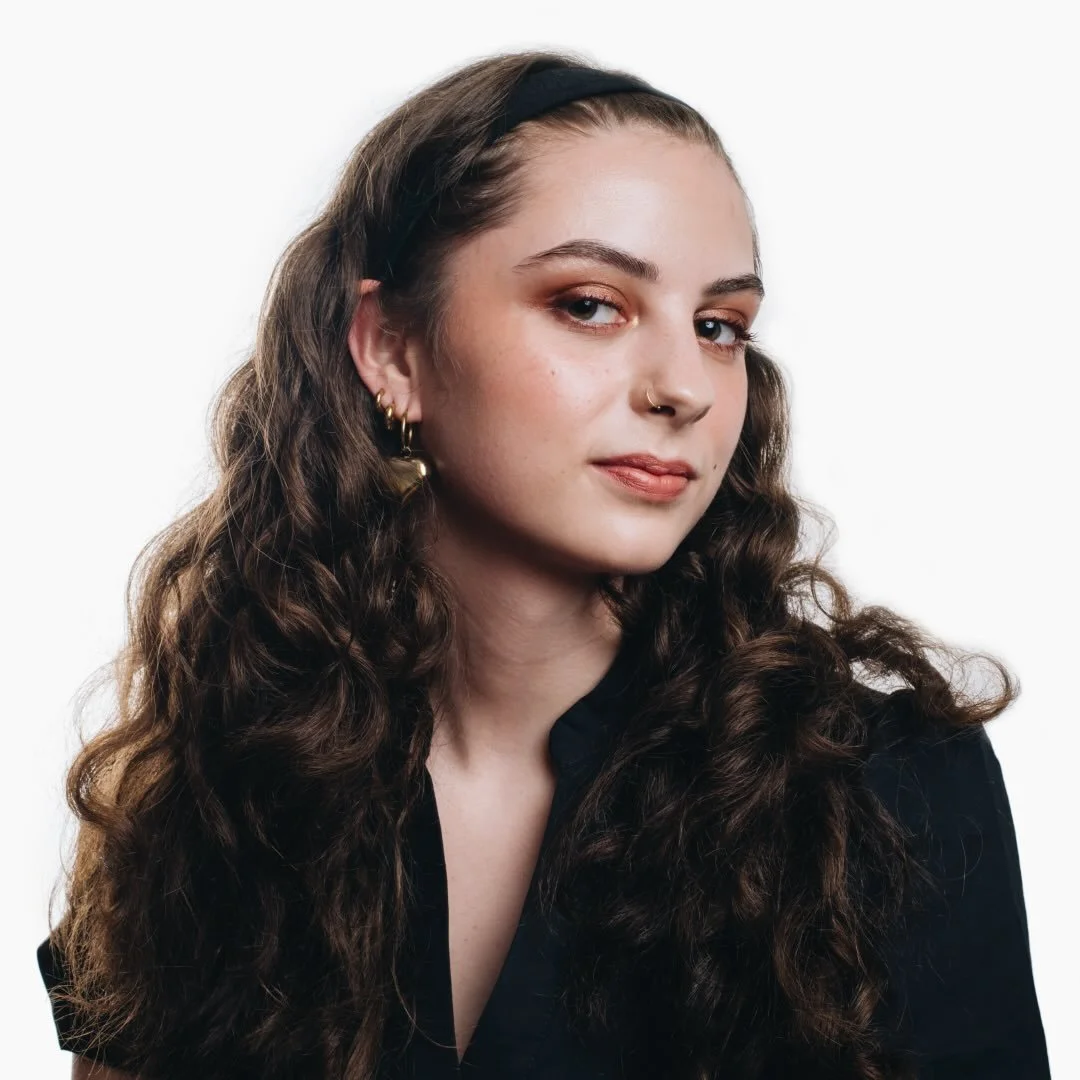 A young woman with long, wavy brown hair, wearing a black headband and a black top, looking at the camera with a slight smile, against a plain white background.