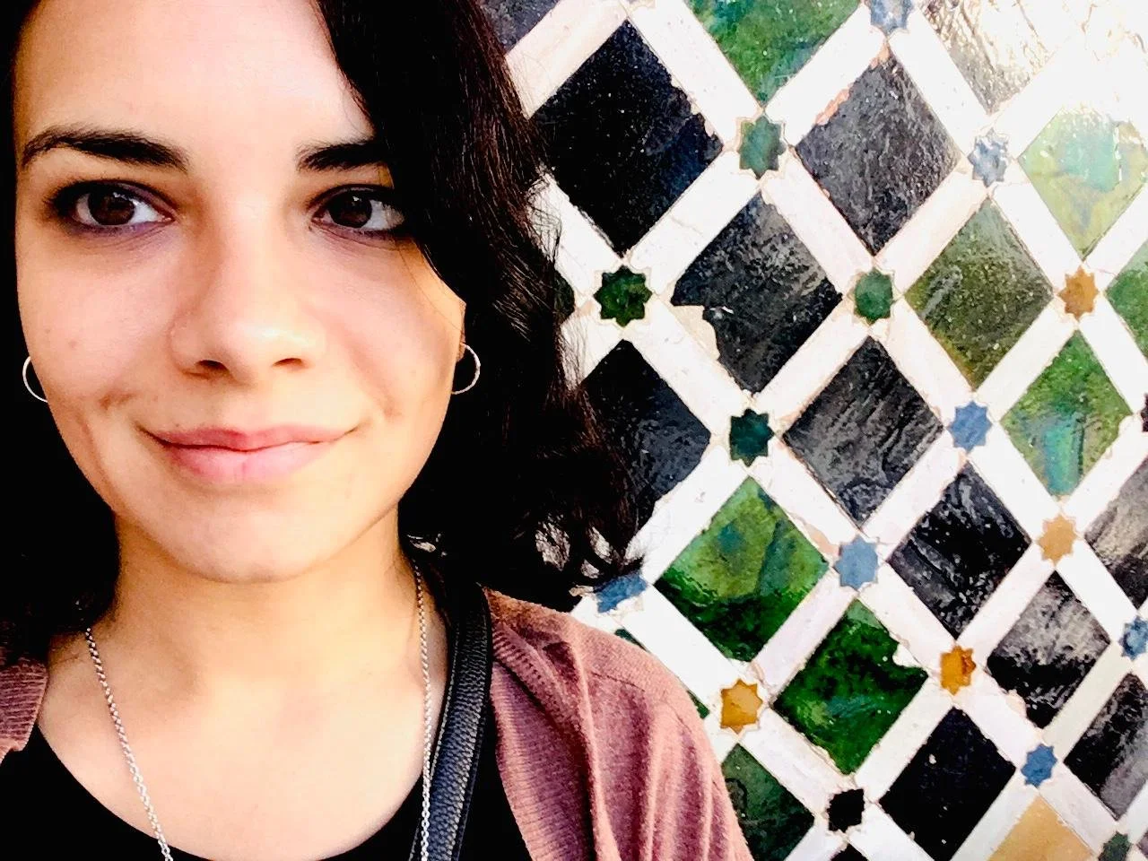 A young woman with dark curly hair, hoop earrings, and a silver necklace taking a selfie outdoors against a decorative tiled wall with colorful diamond-shaped tiles.