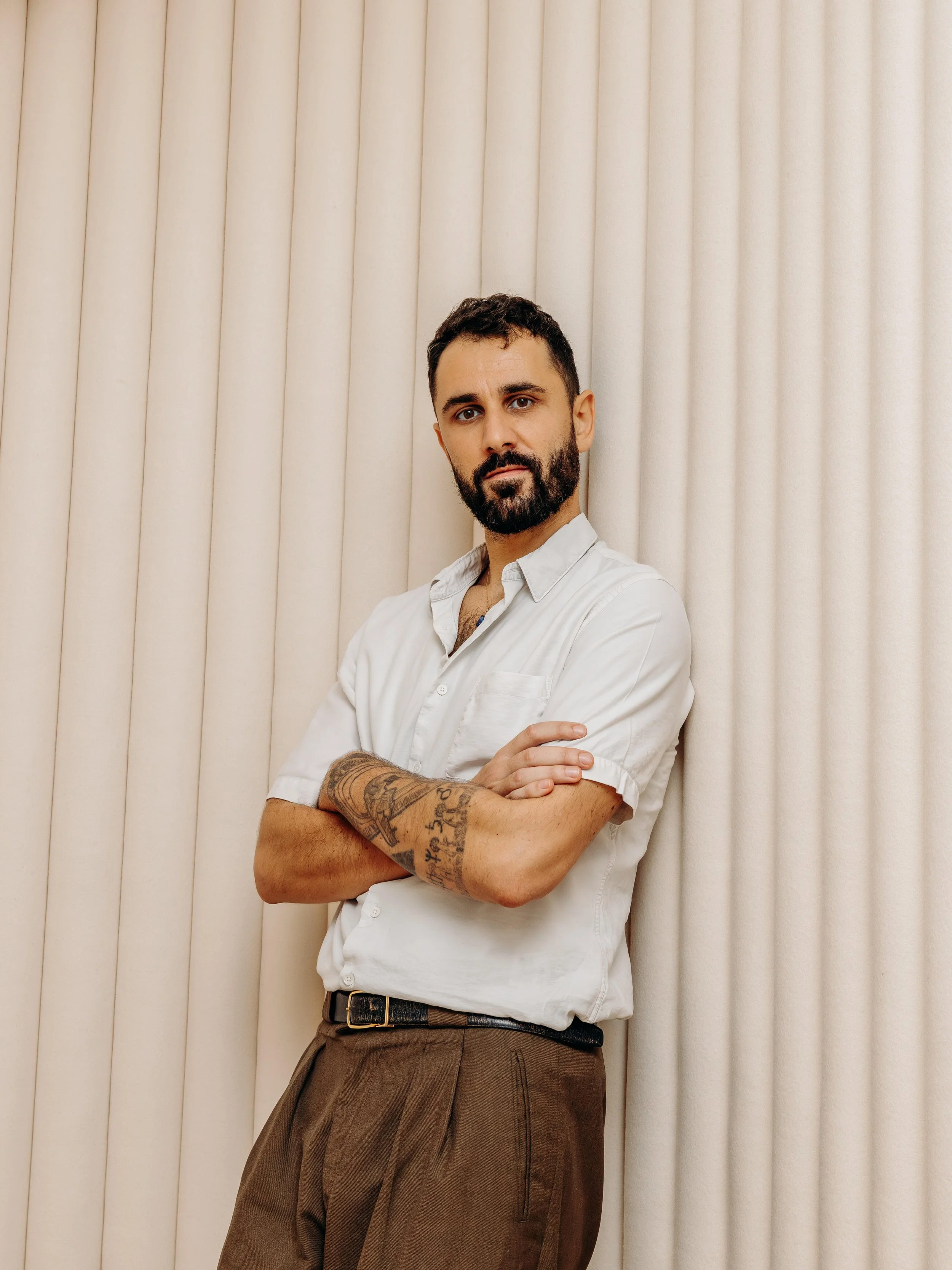 Homme debout, bras croisés, portant une chemise blanche et un pantalon marron, en arrière-plan un mur couleur crème.