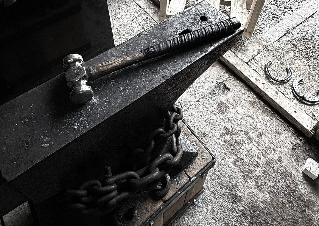 A hammer resting on an anvil with blacksmithing chains and horseshoes nearby.