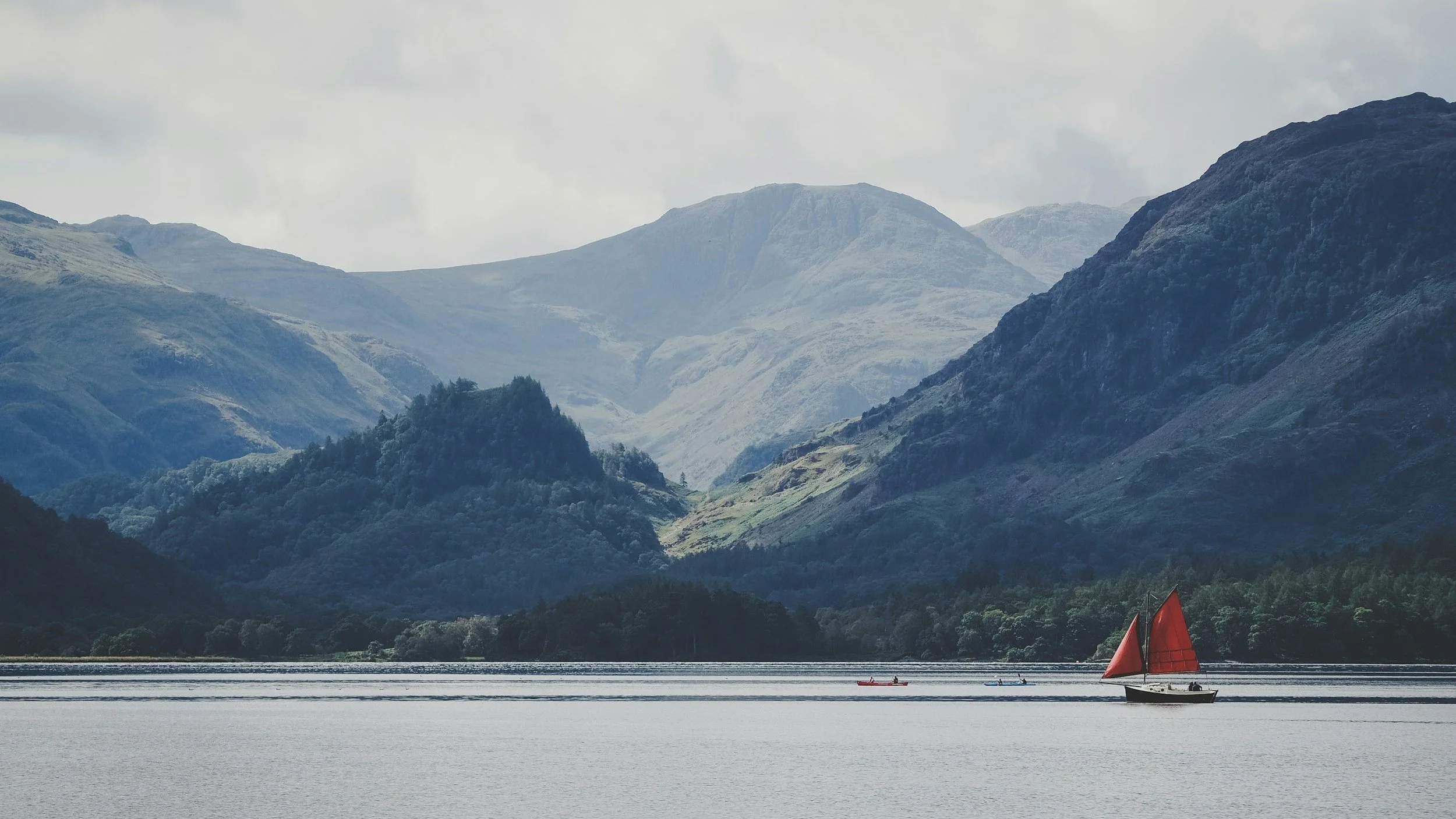 Scenic view of a large lake with a red sailboat and smaller boats, surrounded by green hills and towering mountains in the background, under a partly cloudy sky.