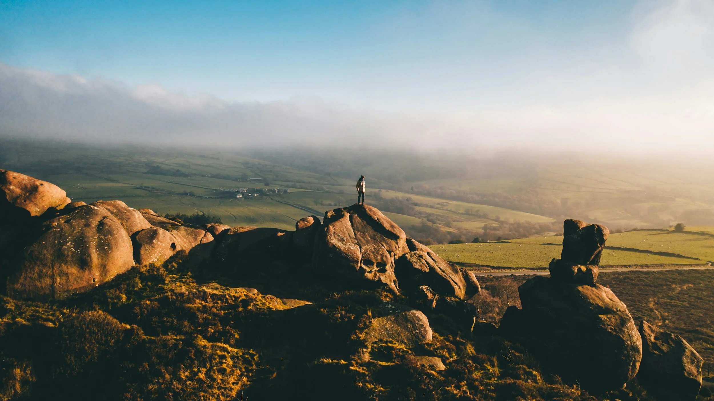 A person standing on rocks overlooking a vast landscape with fields, rolling hills, and a misty horizon during sunset.