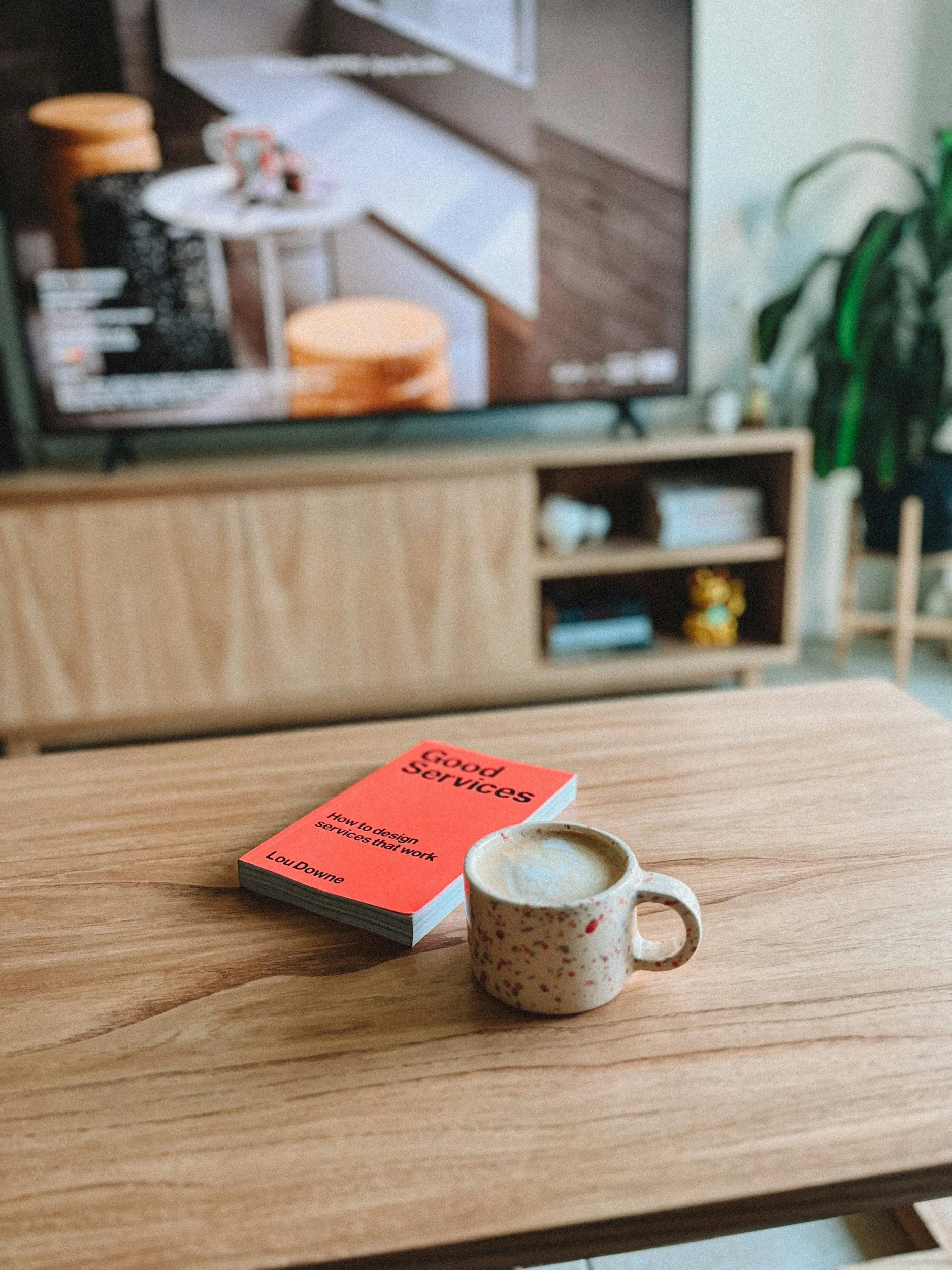 A coffee mug on a wooden table with a book titled 'Good Services' and a coffee cup in front of it, with a TV screen and bookshelf in the background.