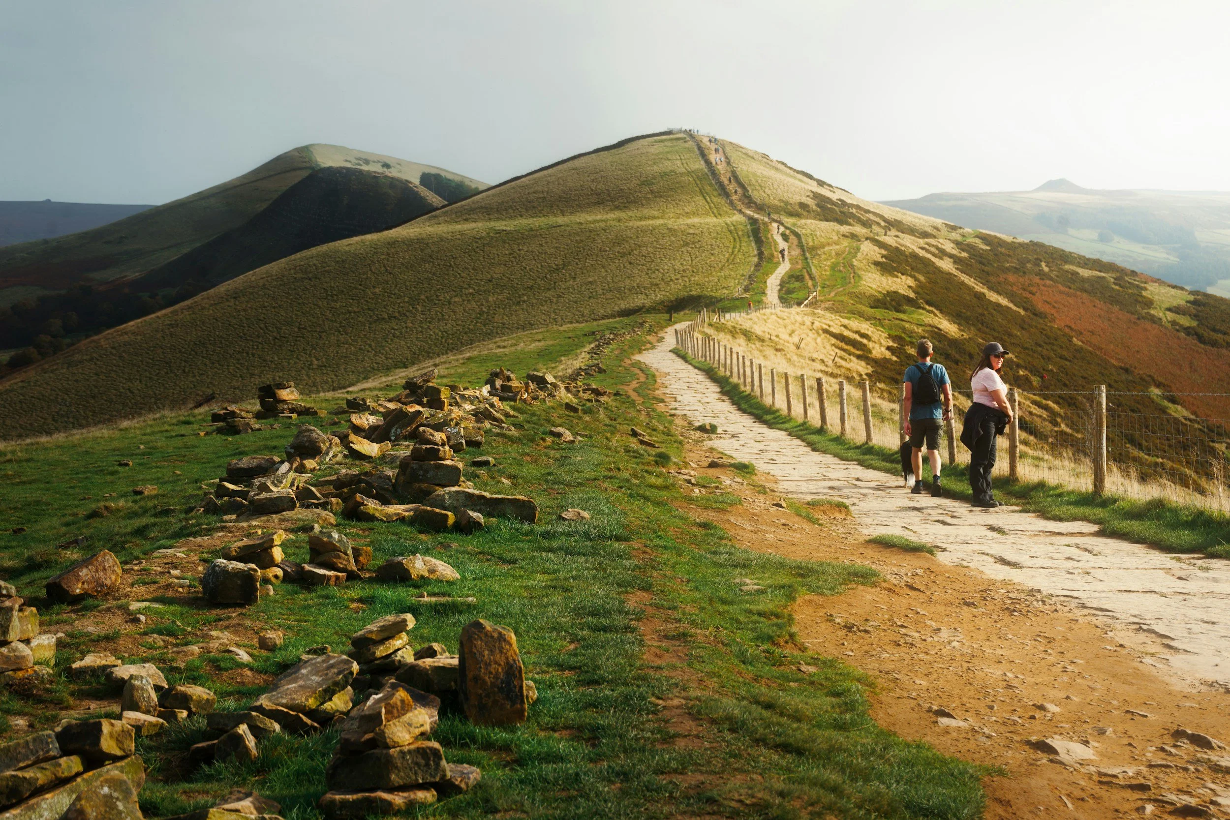 Two hikers walking along a mountain trail with rolling green hills in the background, some rocks near the path, and a fence on the right side.