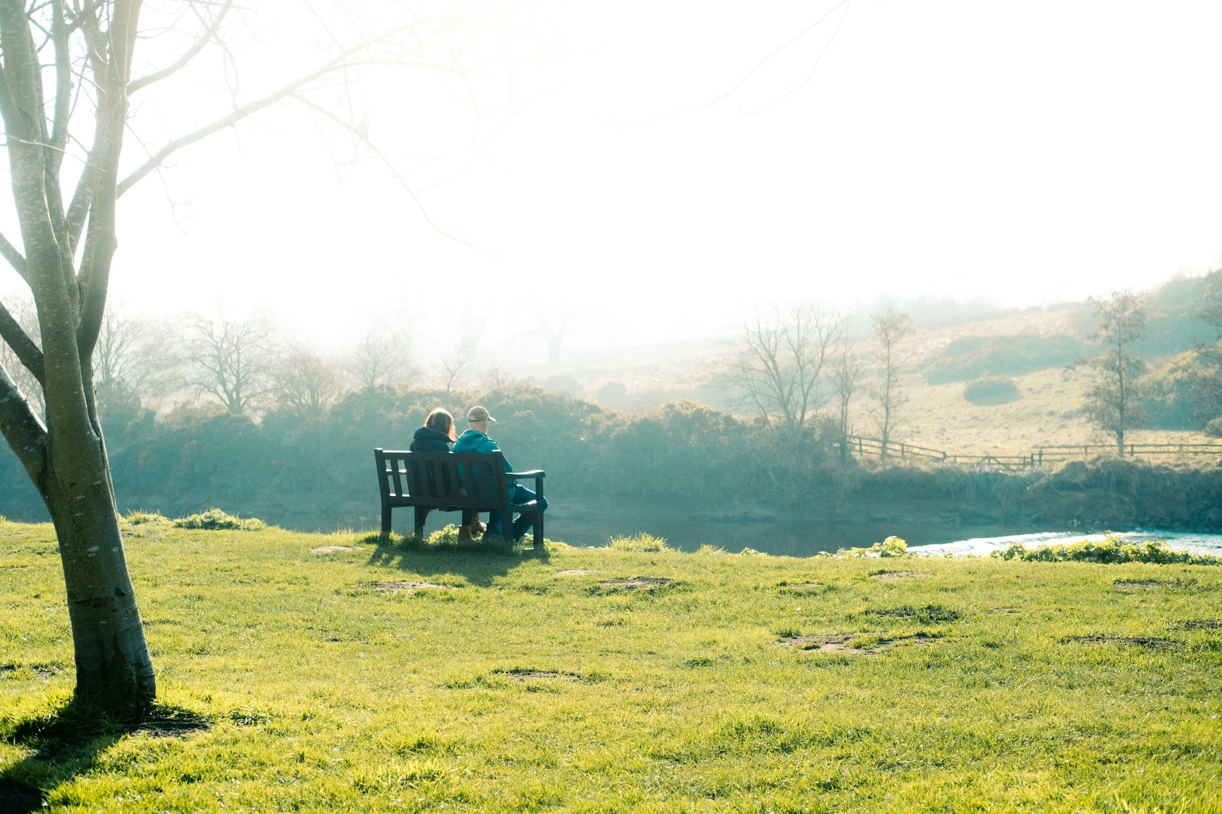 Two people sit on a bench in a grassy park area with trees and hills in the background, during daytime with bright sunlight.