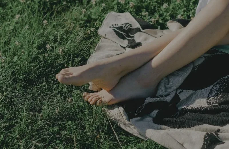 Person sitting outdoors on grass, with legs extended and feet visible, on a patterned blanket.