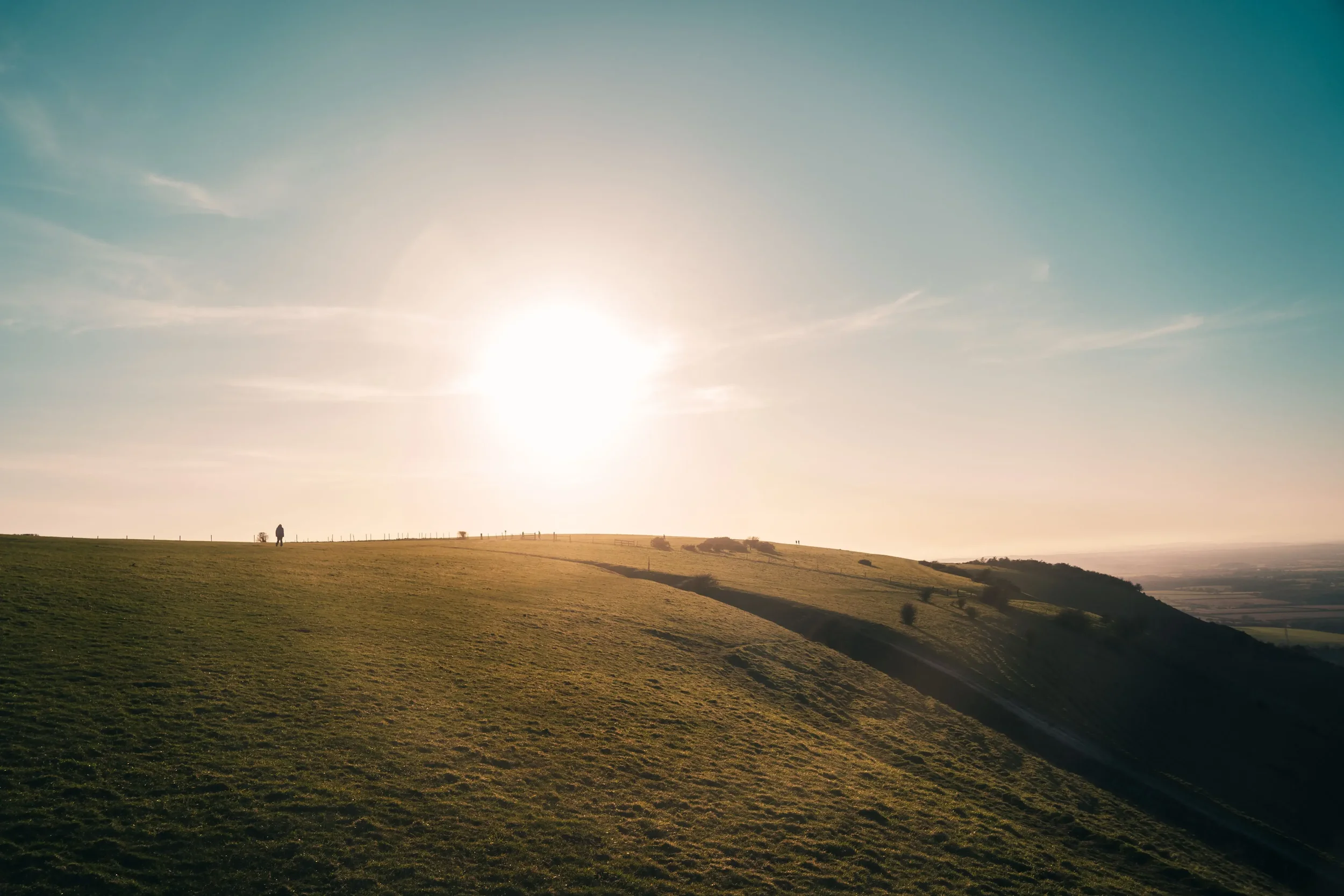A landscape of rolling green hills at sunset with a person walking in the distance and a few scattered trees and bushes.