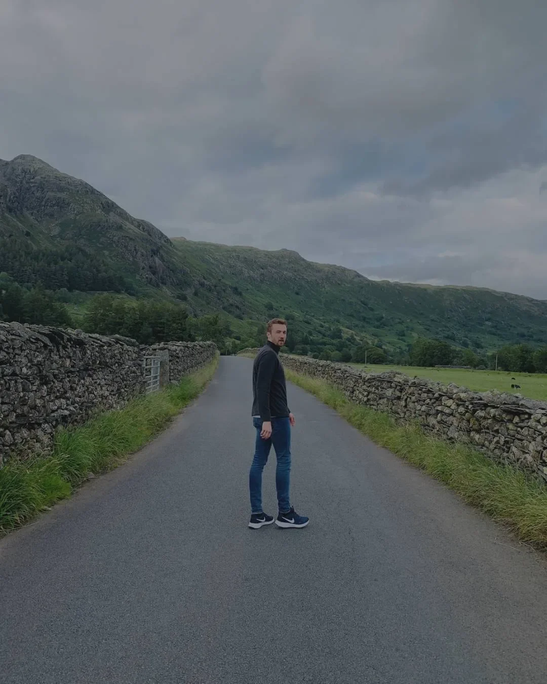 A man standing in the middle of a rural road bordered by stone walls, with lush green hills and mountains in the background under a cloudy sky.