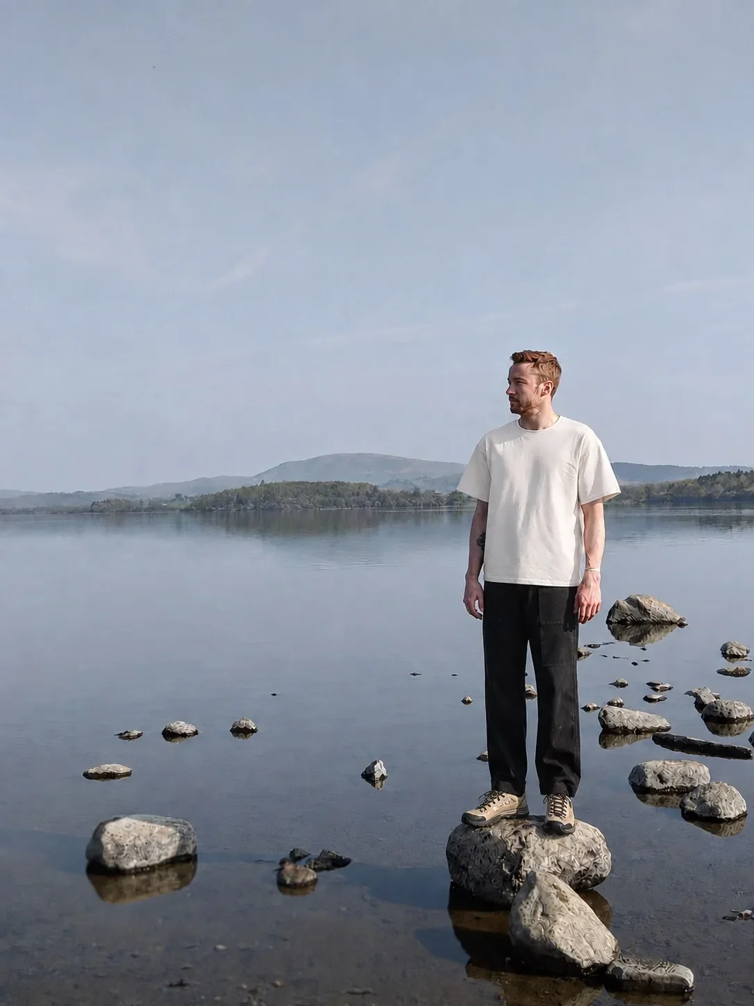 A man standing on a rock in a calm lake with mountains in the background, wearing a white t-shirt and black pants.