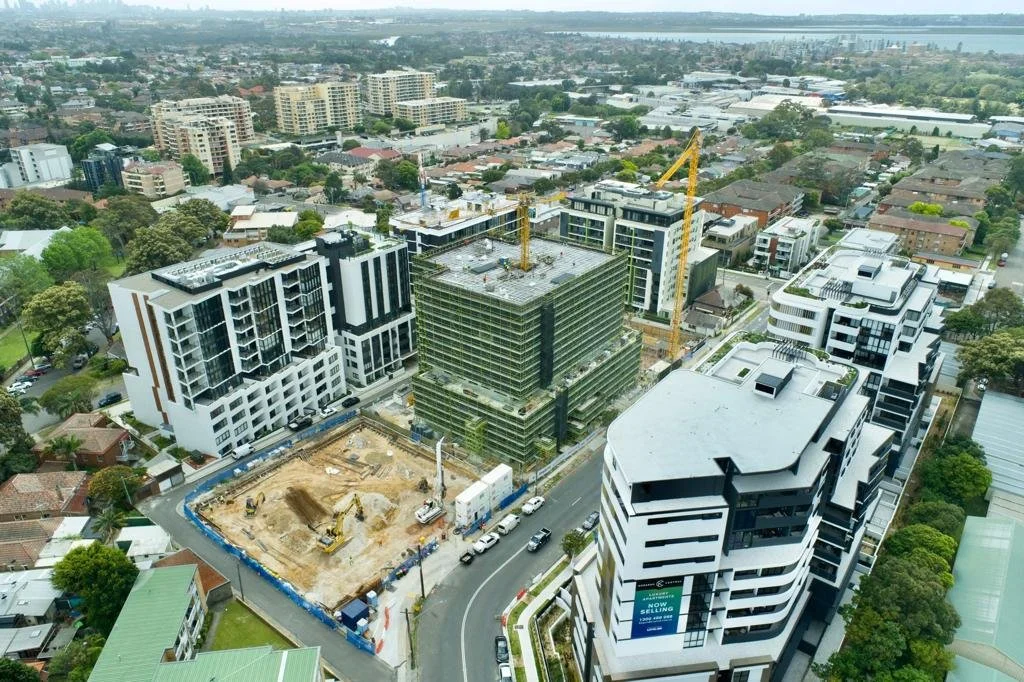 An aerial view of a construction site surrounded by modern apartment buildings and residential houses, with cranes and construction equipment visible, in a city with a river in the background.