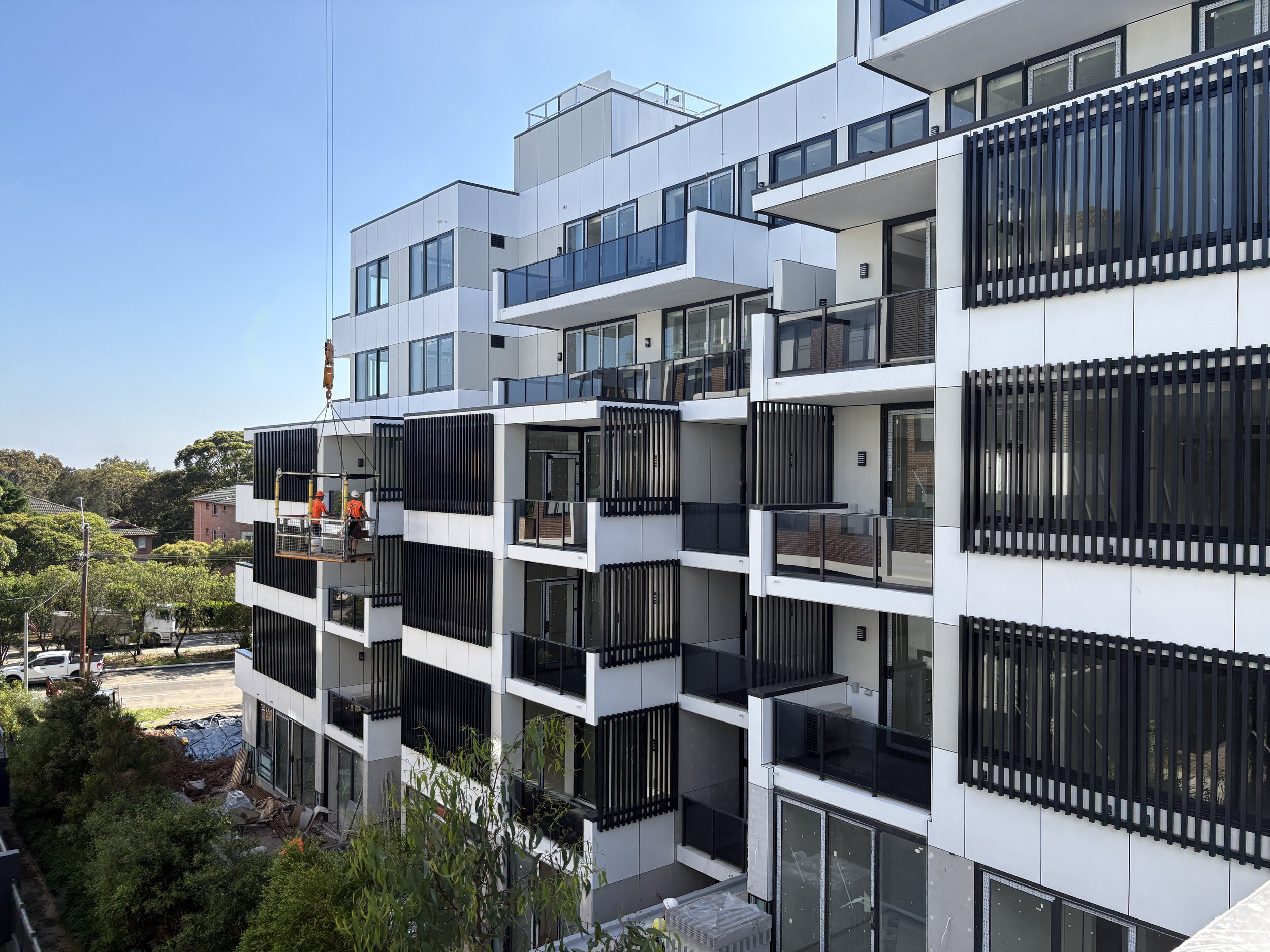 A modern multi-story apartment building with black balcony railings and large windows, under construction with workers on a lift, set against a clear blue sky.