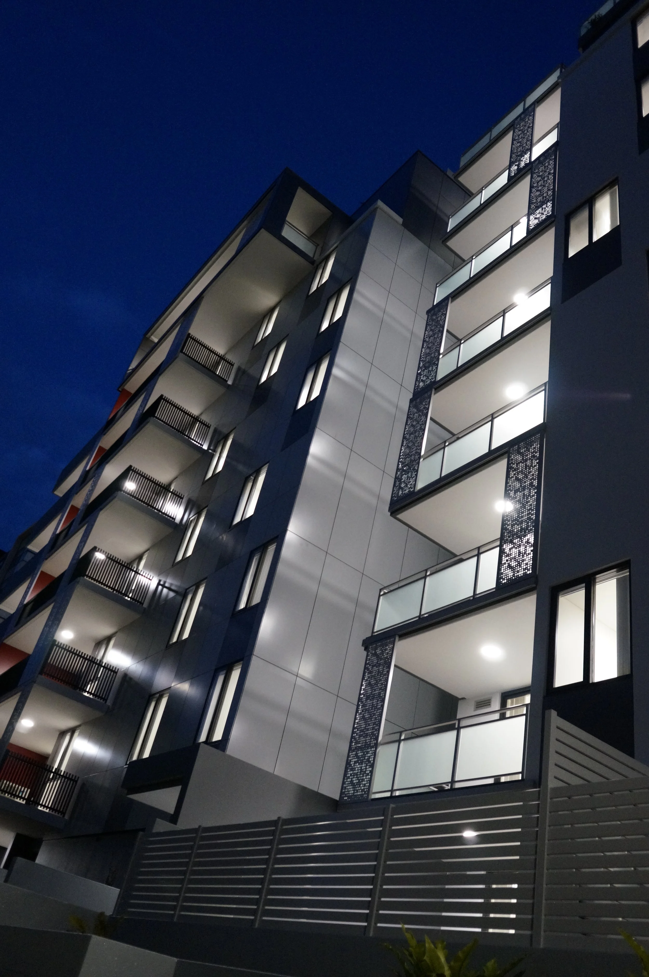 Modern apartment building at night with illuminated windows and balconies, dark exterior, and a deep blue evening sky.
