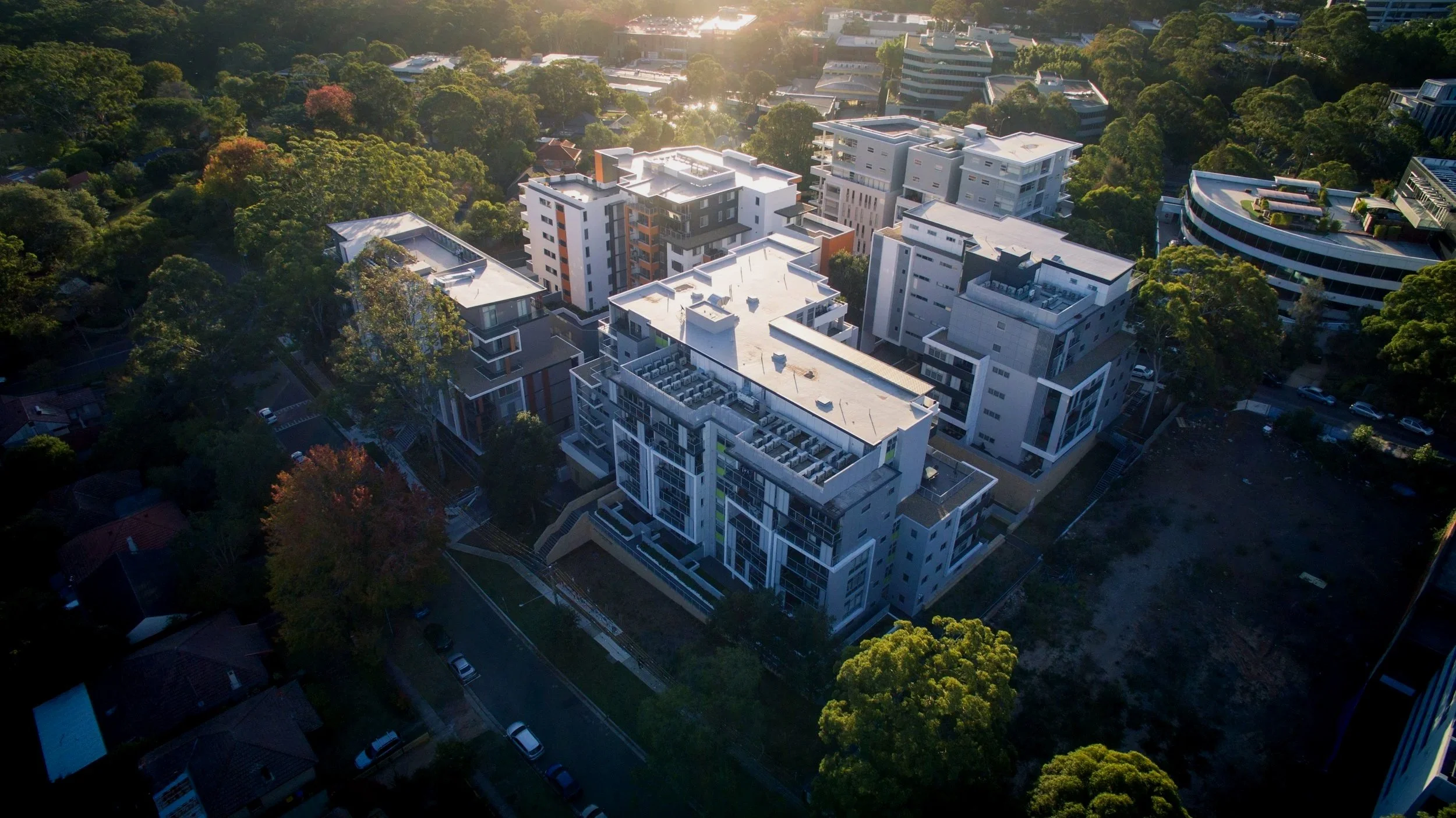 Aerial view of a modern multi-story apartment complex surrounded by green trees, with sunlight reflecting off the rooftops in a suburban area.