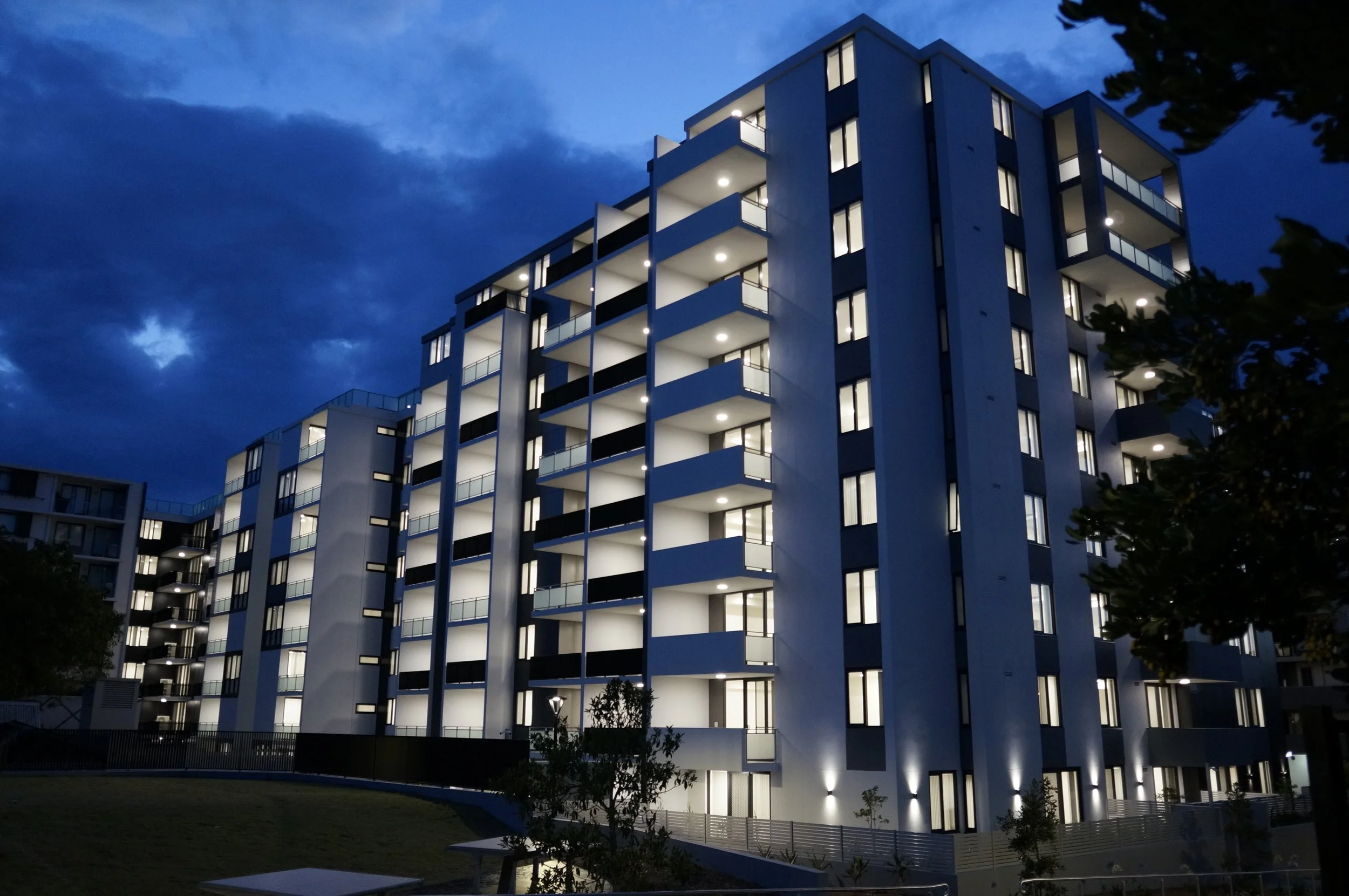 Nighttime view of a modern multi-story apartment building with illuminated windows and balconies, surrounded by trees and a grassy area.