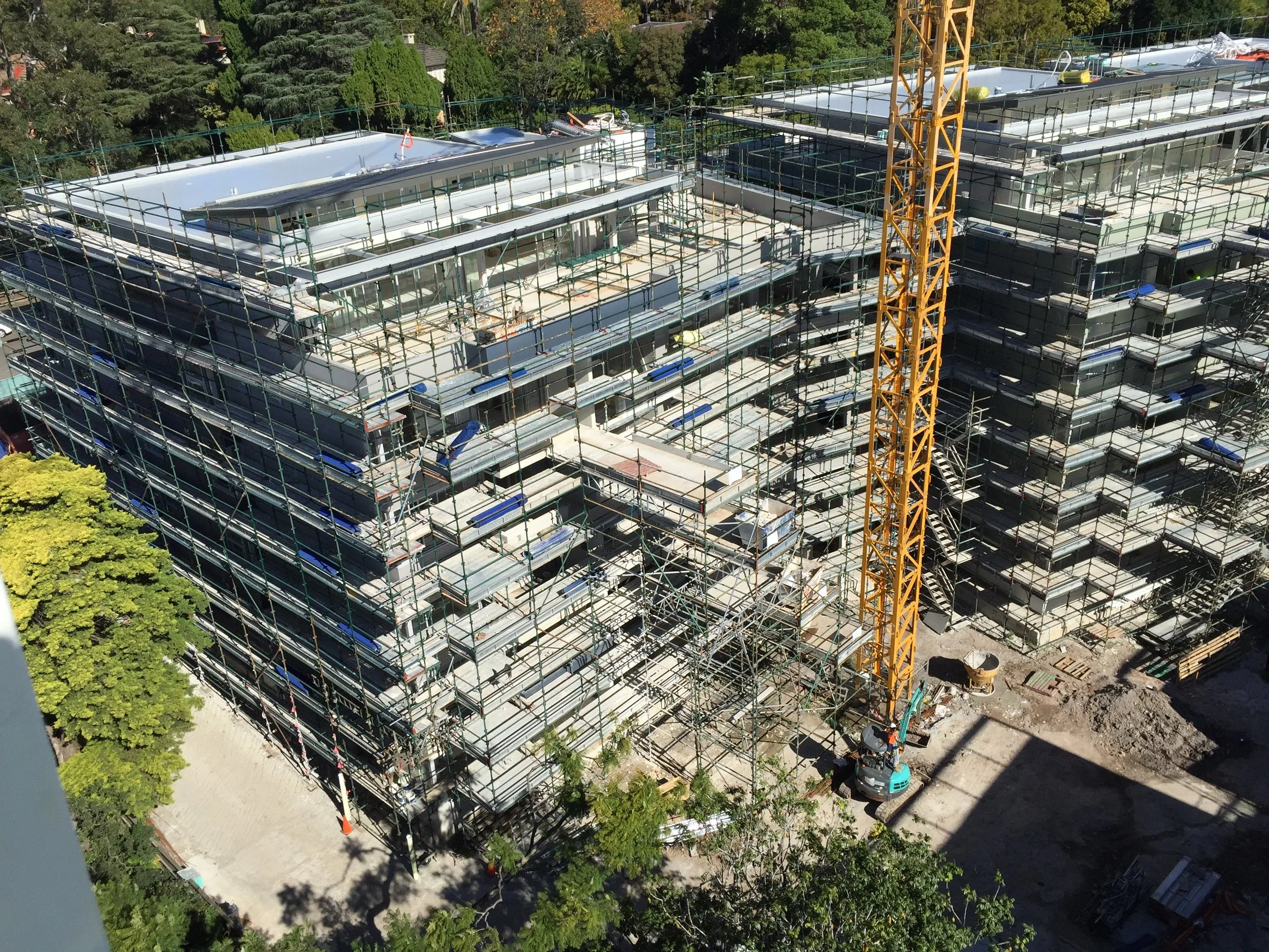 A high-angle view of a building under construction with scaffolding, a tower crane, and construction workers. There are trees surrounding the site and construction materials on the ground.