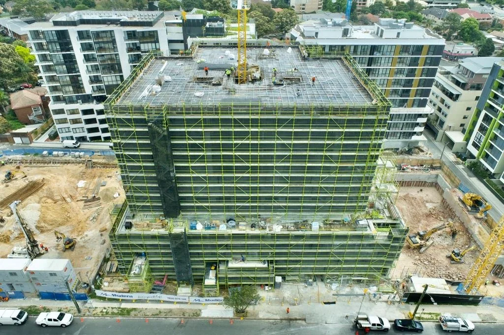 A high-rise building under construction with workers on the roof, surrounded by scaffolding, cranes, and construction equipment, in an urban area.