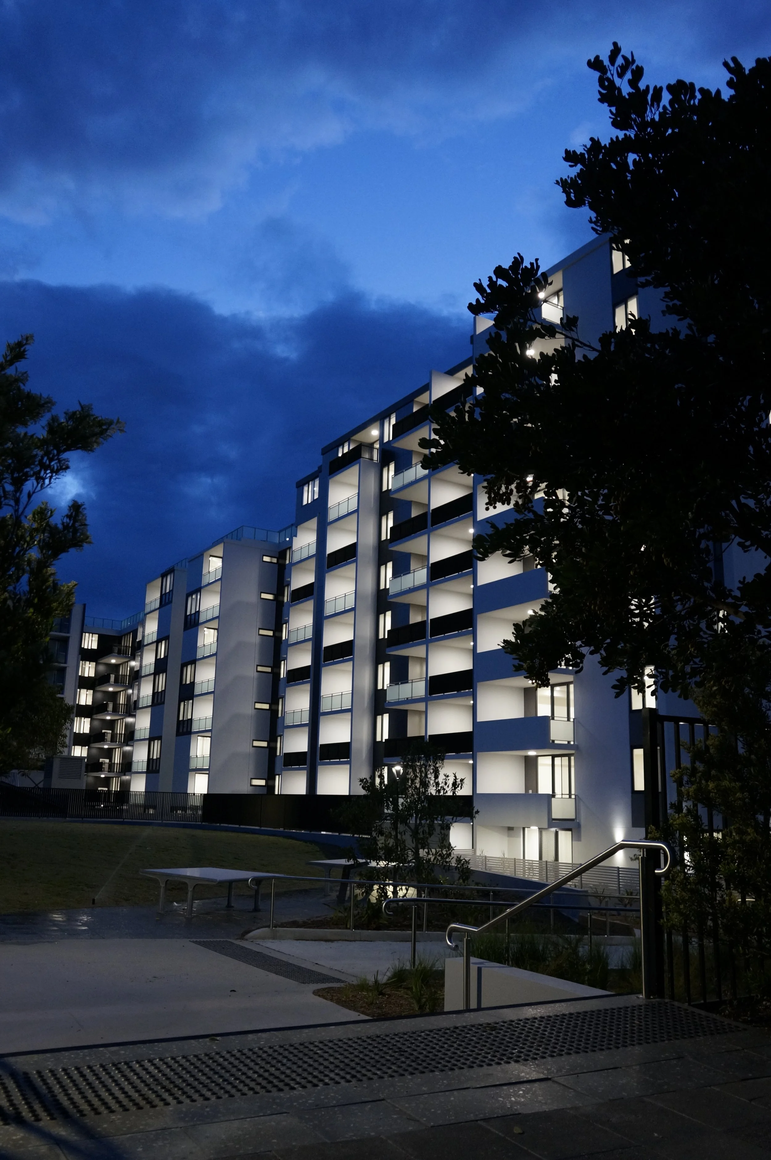 Nighttime view of a modern white multi-story apartment building with lit windows, surrounded by trees, under a partly cloudy blue sky, with outdoor benches and railings in the foreground.