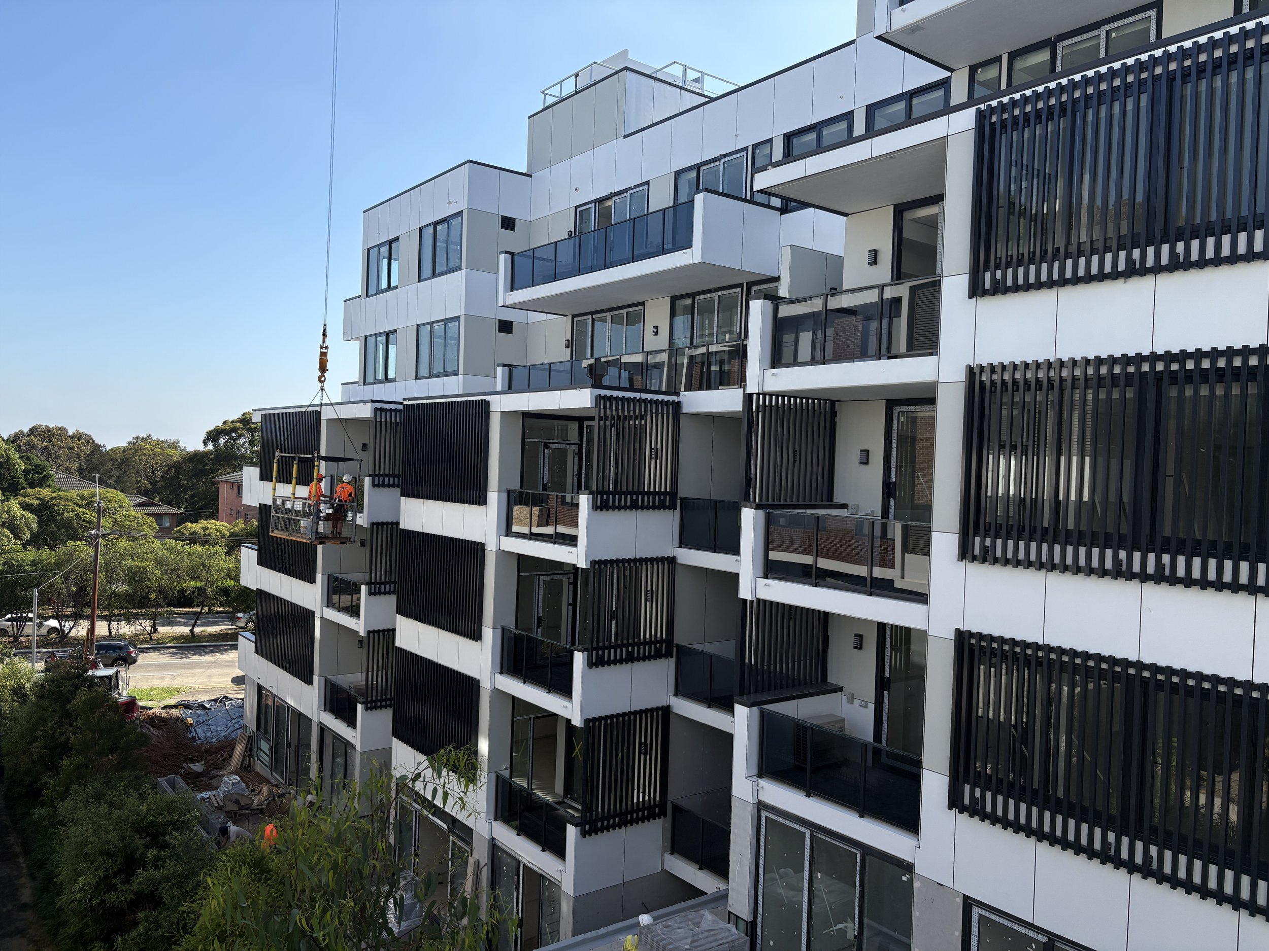 Modern multi-story residential building under construction with workers on scaffolding, surrounded by trees and parked cars.