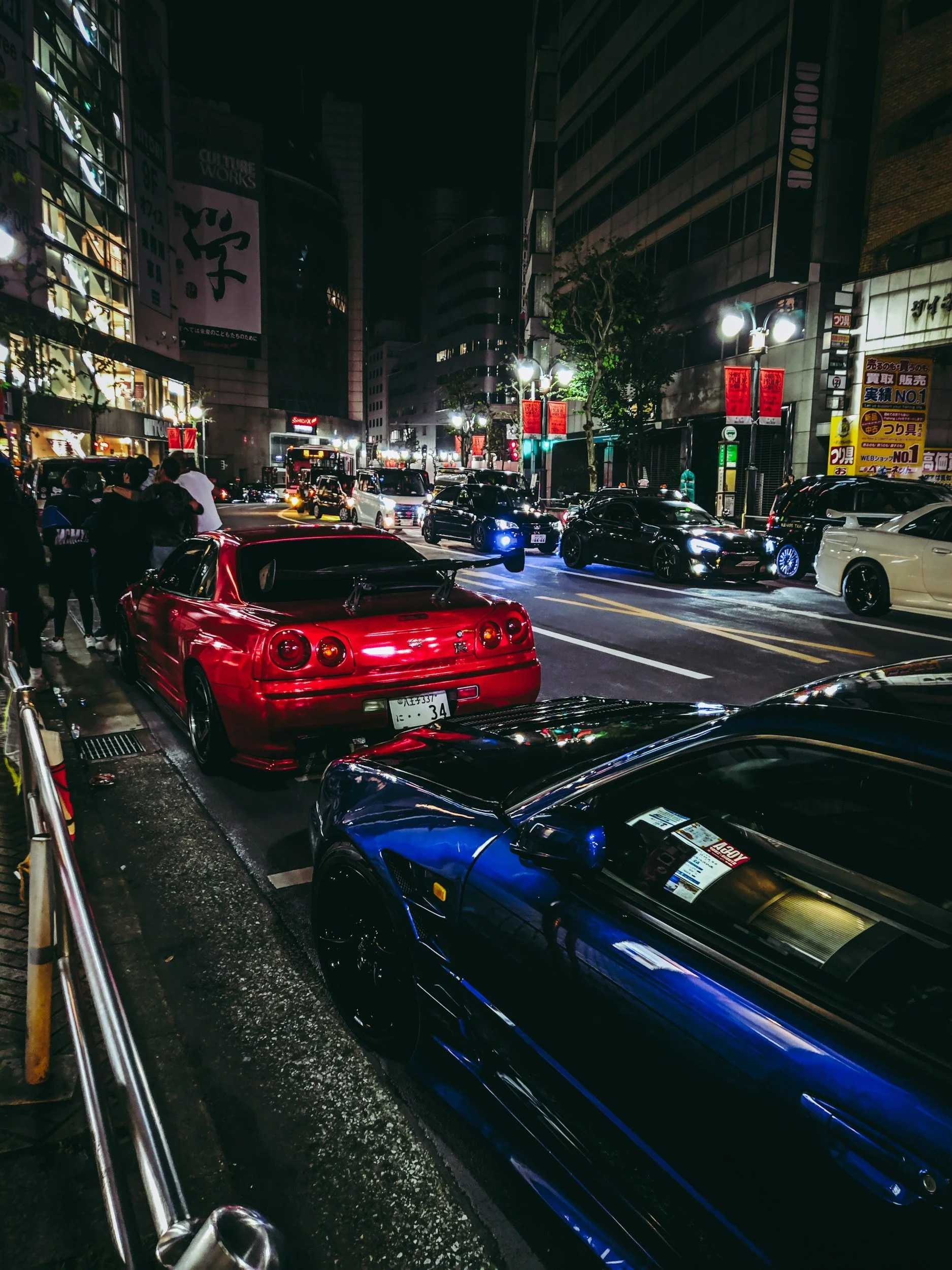 Nighttime city street scene with parked sports cars, including a red car in the foreground and a blue car nearby, along with several other cars and a group of people on the sidewalk.