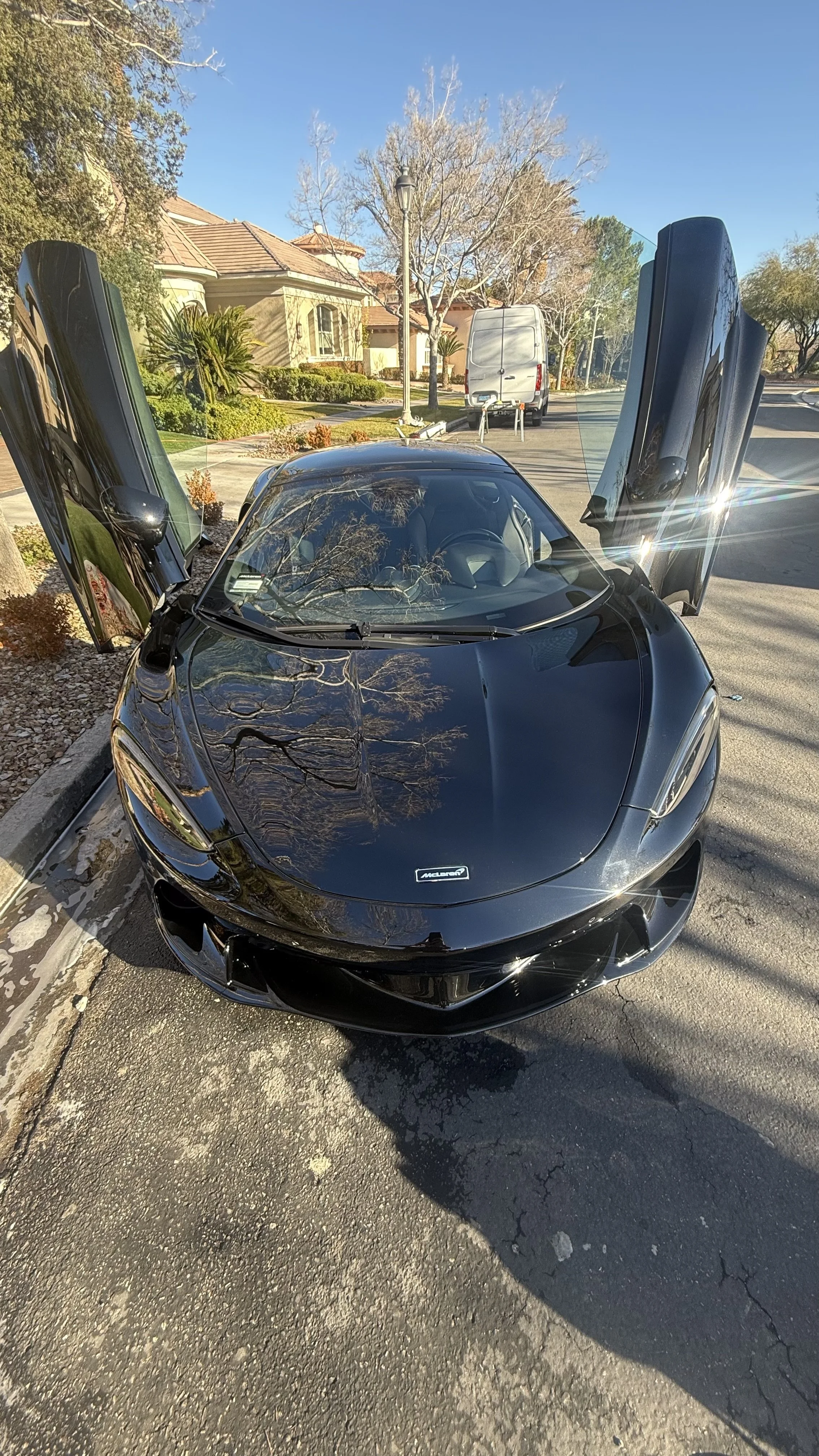 A clean black McLaren sports car with its dihedral doors open parked on a residential street, with reflections of trees on its shiny surface and houses in the background. Cool cars Las Vegas.