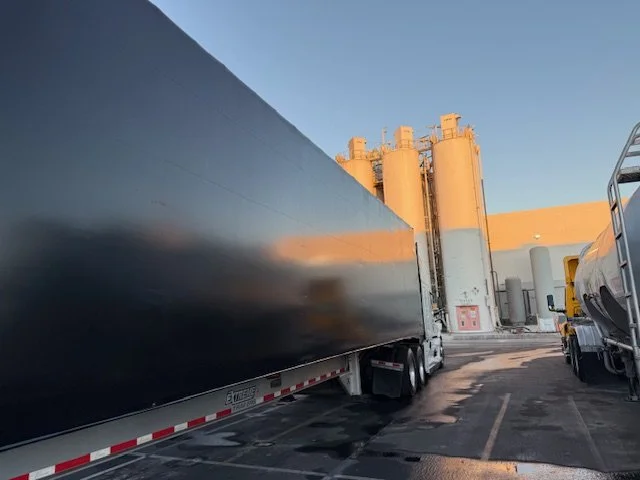 Large black semi-truck Conestoga trailer parked in an industrial area near silos, with another truck visible to the right, under a clear blue sky in the late afternoon. Rainx, alkaline, hand wash. commercial vehicle cleaning.