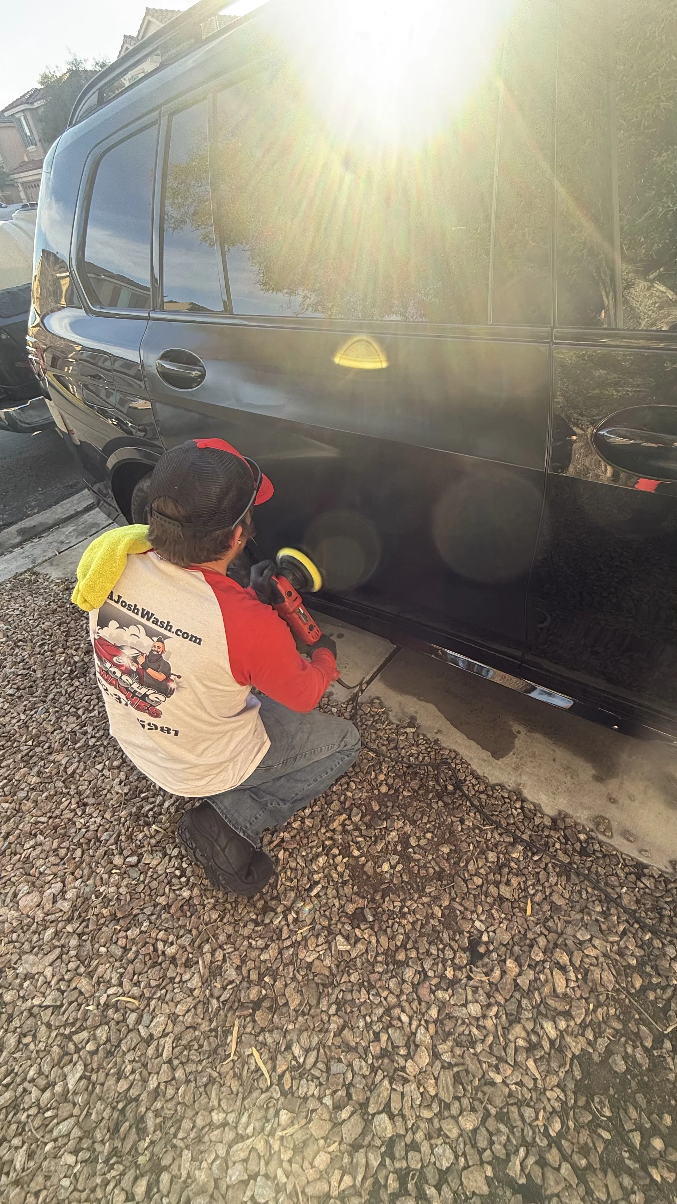 A man is crouched down washing or polishing a black car with a power buffer, wearing a beige and red shirt with a logo and website, a black cap with red accents, and black shoes, on a gravel driveway with houses in the background and bright sunlight.