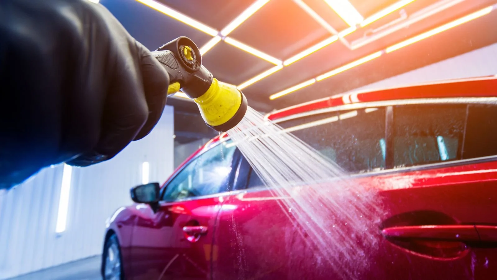 A person washing a red car with a yellow and black hose in an indoor wash bay with bright, linear ceiling lights.