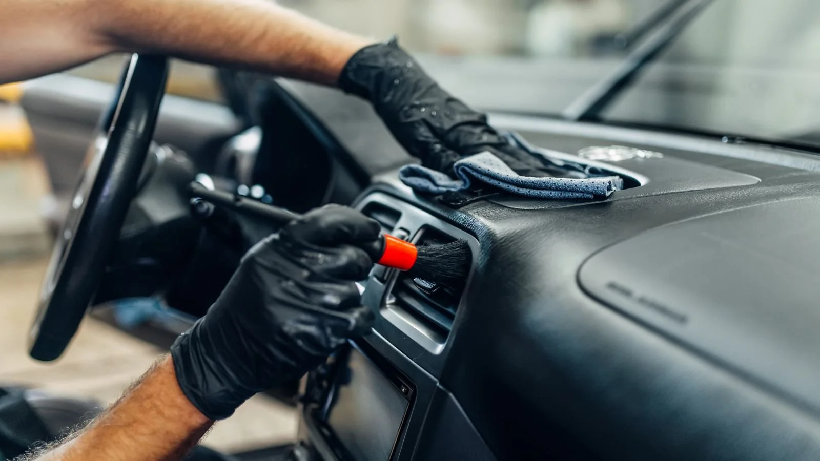 A person wearing black gloves cleaning the interior dashboard of a vehicle using a brush and cloth. Deep interior cleaning of car. Delicate professional care.