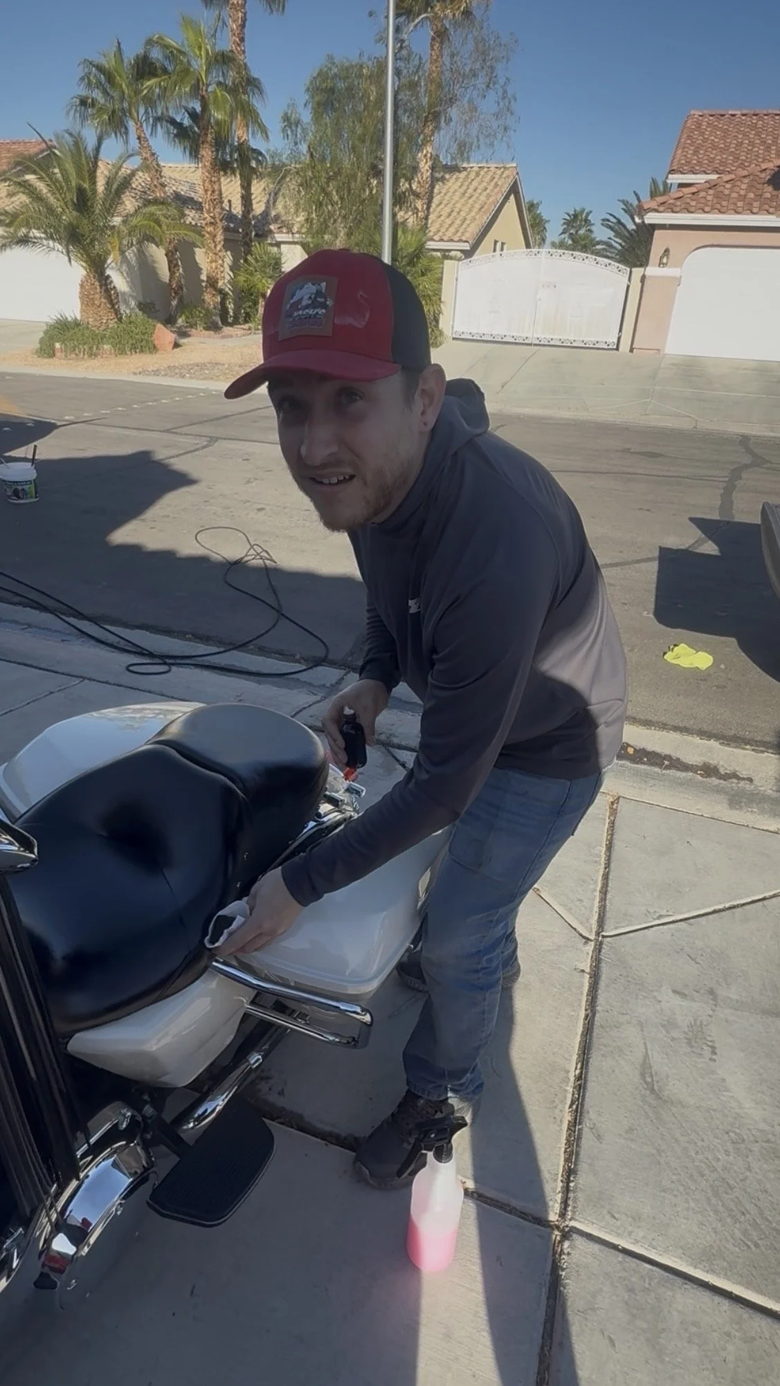 A man wearing a red and black baseball cap and gray jacket is cleaning a white motorcycle outside on a sunny day. Houses with red-tiled roofs and palm trees are visible in the background.