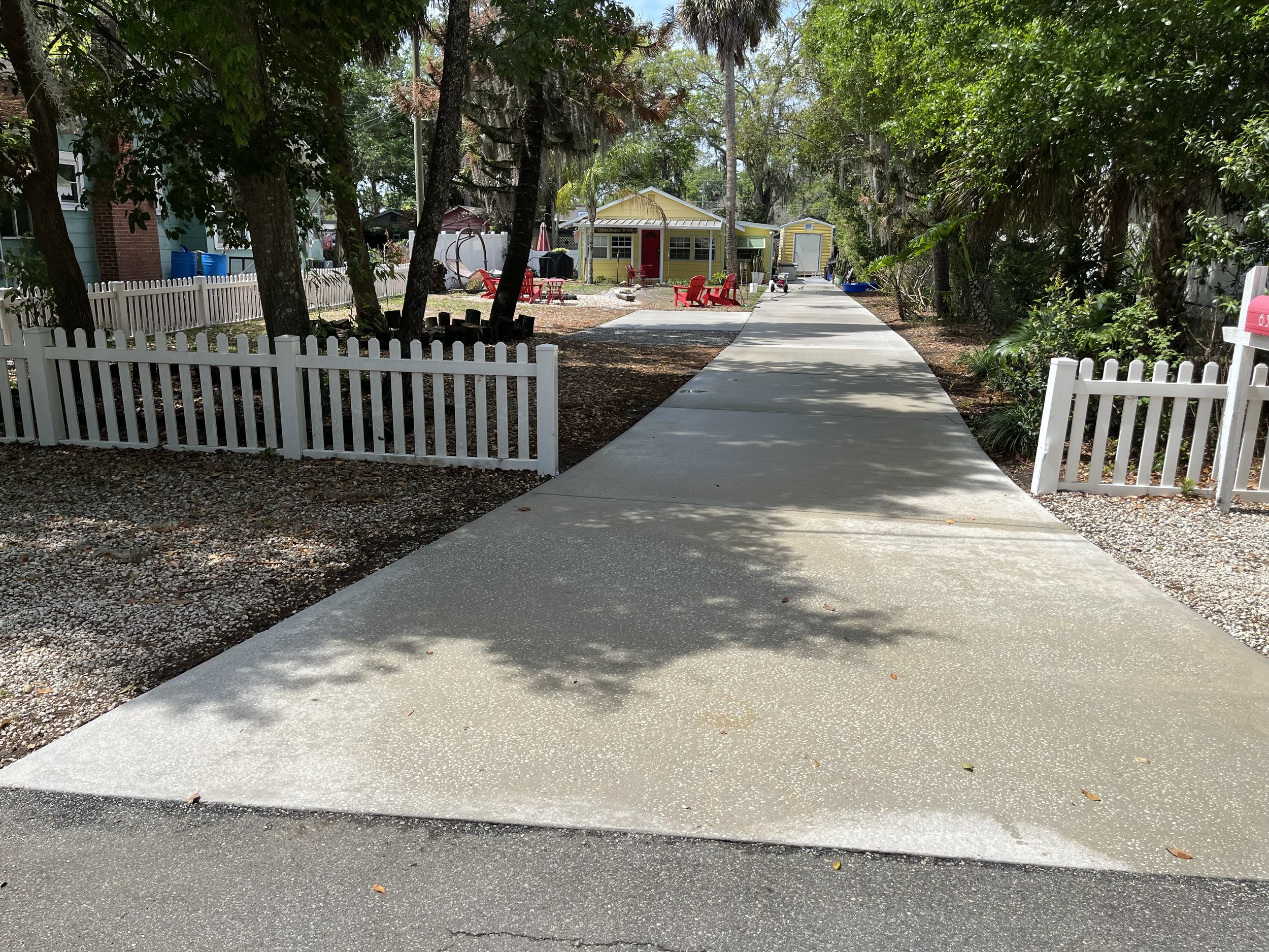 A sidewalk in a residential neighborhood with trees on either side. In the background, there are small colorful houses, one yellow with a red door and another yellow with a white garage, along with outdoor furniture and play equipment. A white picket