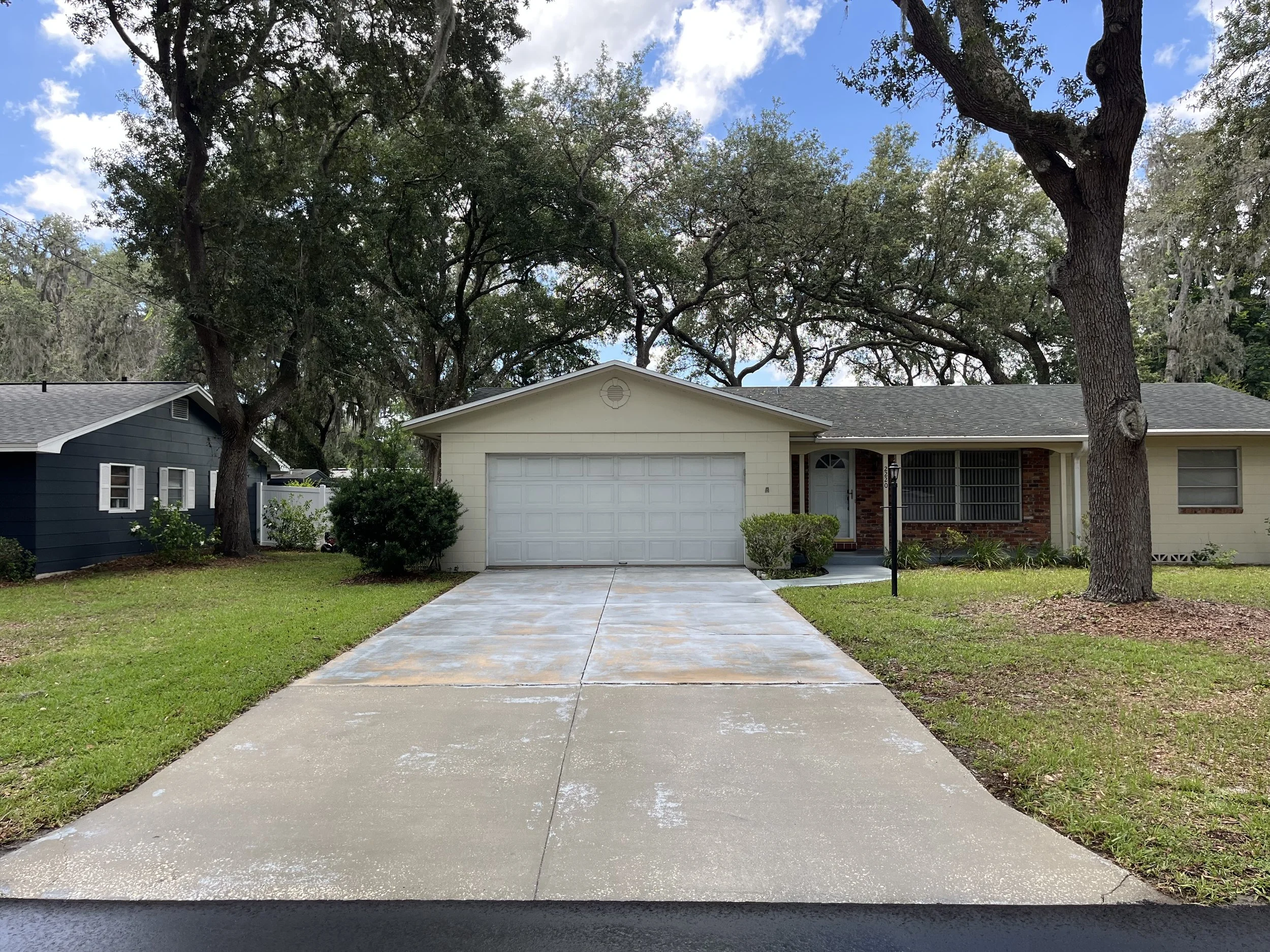 front yard of a suburban house with driveway, lush green lawn, large trees, and a partly cloudy sky