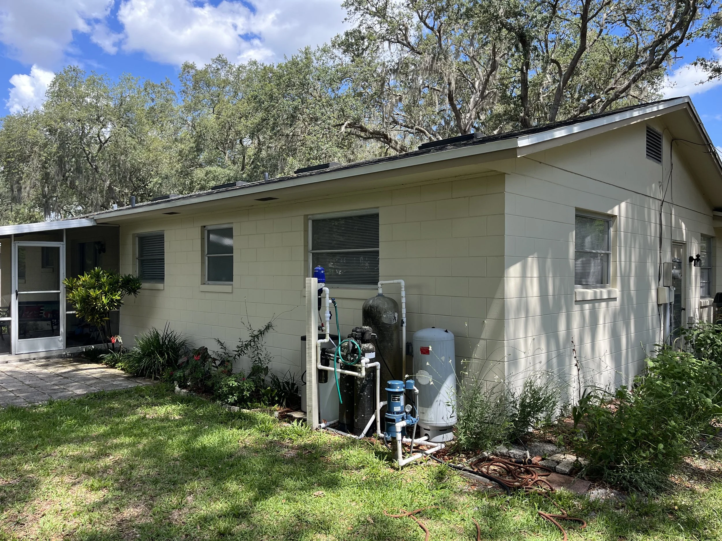 The back side of a house with a yellow exterior, multiple windows, and a sloped roof with solar panels. There is a small patio area with plants, garden hoses, and water filtration equipment outside.