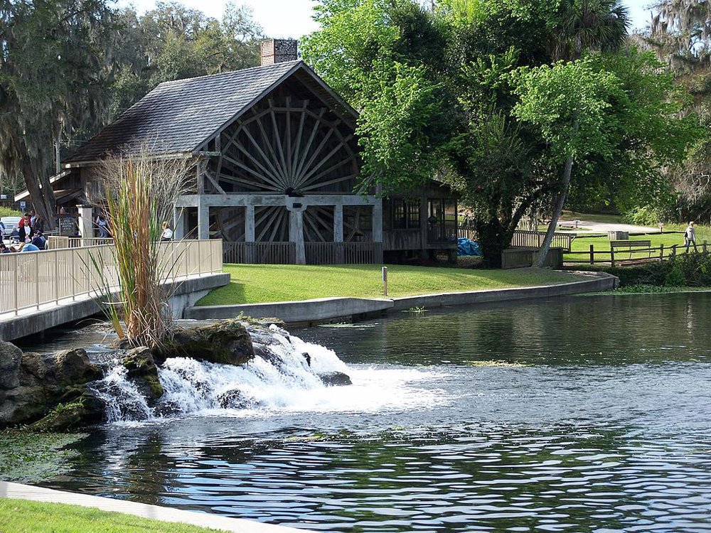 A park scene with a water mill building, a pond, and a small waterfall with surrounding trees and grass.
