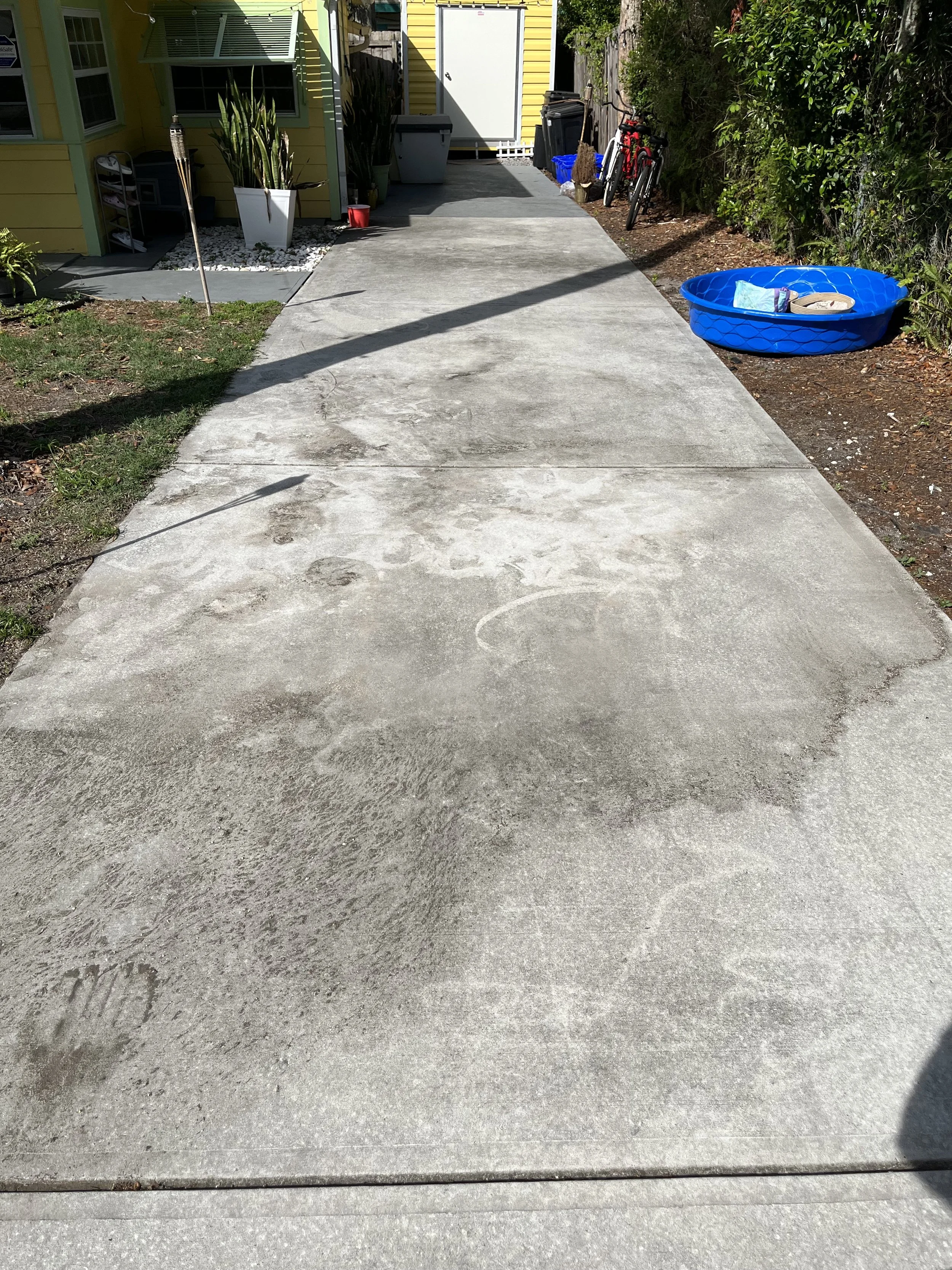 Concrete pathway leading to a yellow house with green trim, with plants, bicycles, and a blue plastic pool with items inside on the right side.