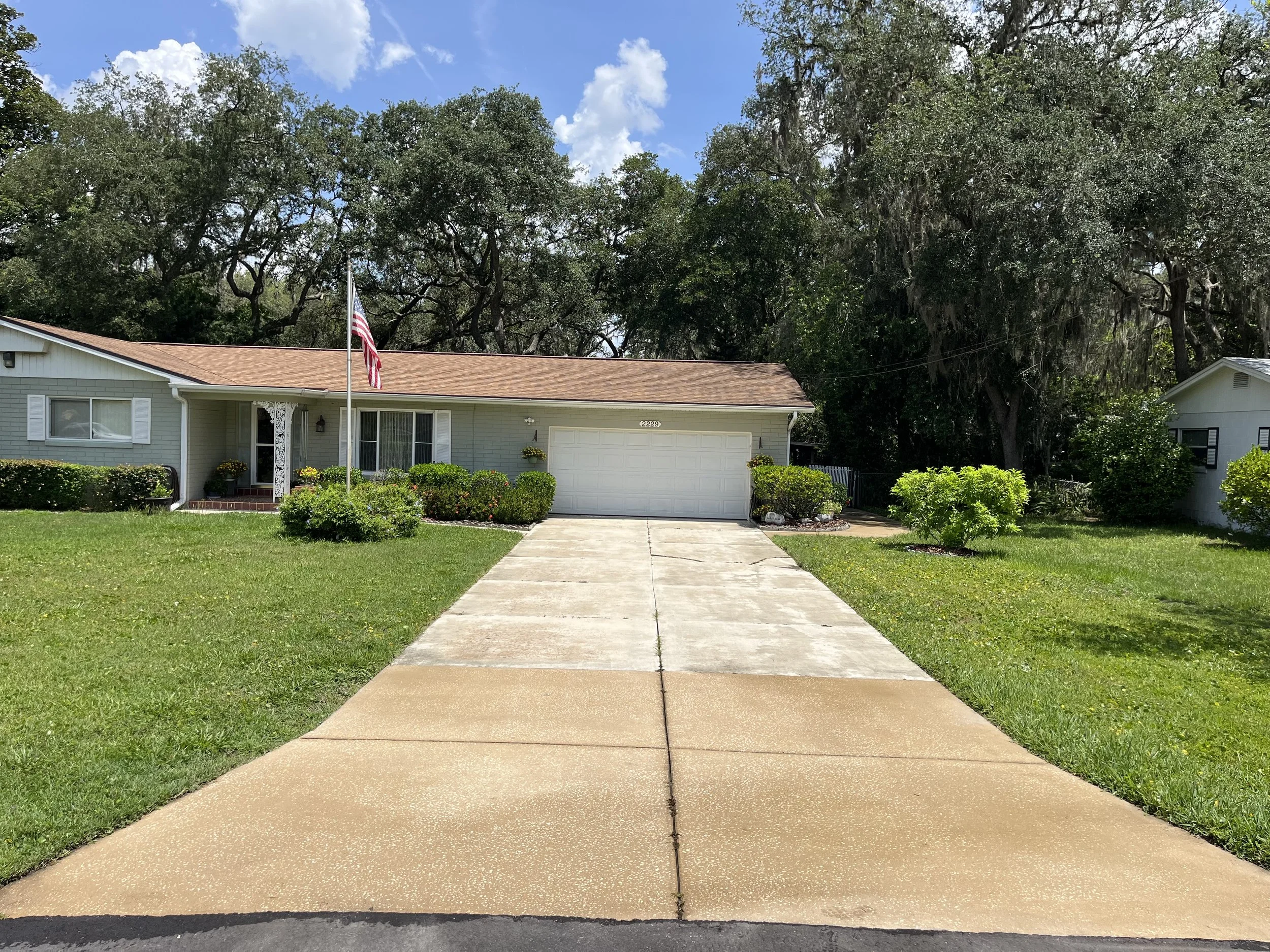 Front view of a single-story house with a light gray exterior, white shutters, and a brown shingle roof. An American flag is mounted on a pole in the front yard. The house number 2229 is visible on the garage door. The driveway is concrete leading to