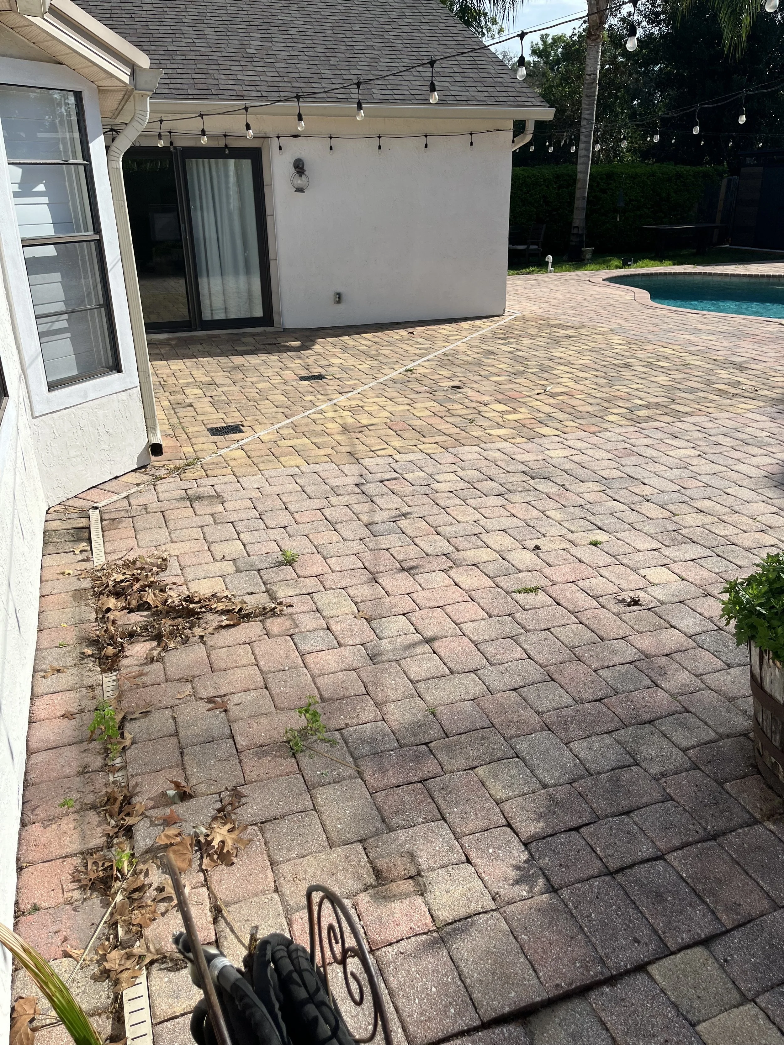 Backyard with brick patio, string lights, a section of swimming pool, and some fallen leaves near the house wall. Part of a stroller is visible at the bottom of the image.